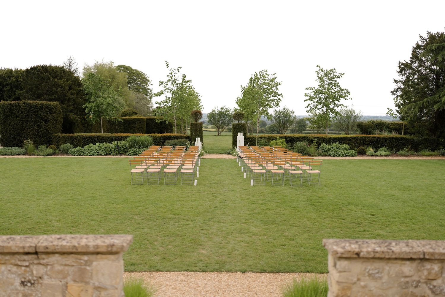 Elegant outdoor wedding ceremony setup with rows of wooden chairs on a manicured lawn surrounded by hedges and trees.