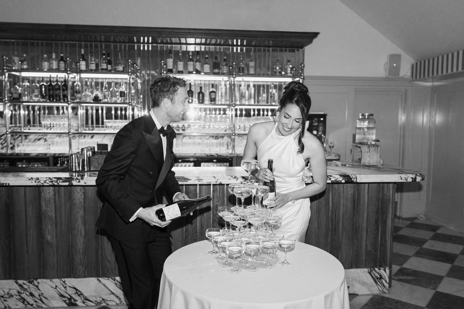 Black and white photo of couple building champagne tower at wedding reception bar.