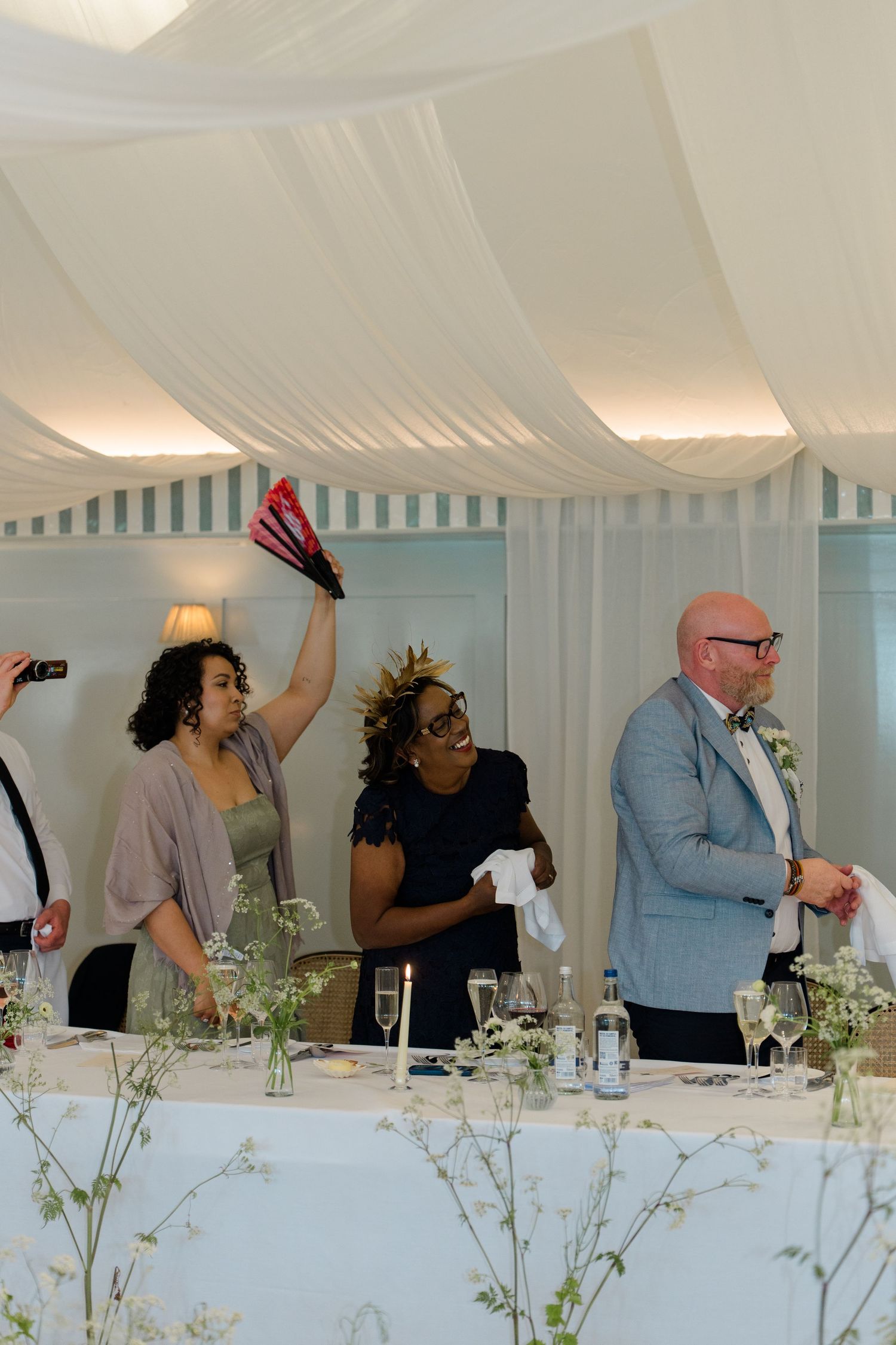 Wedding guests celebrating inside a marquee, woman raising red fans while others laugh and clap at reception.