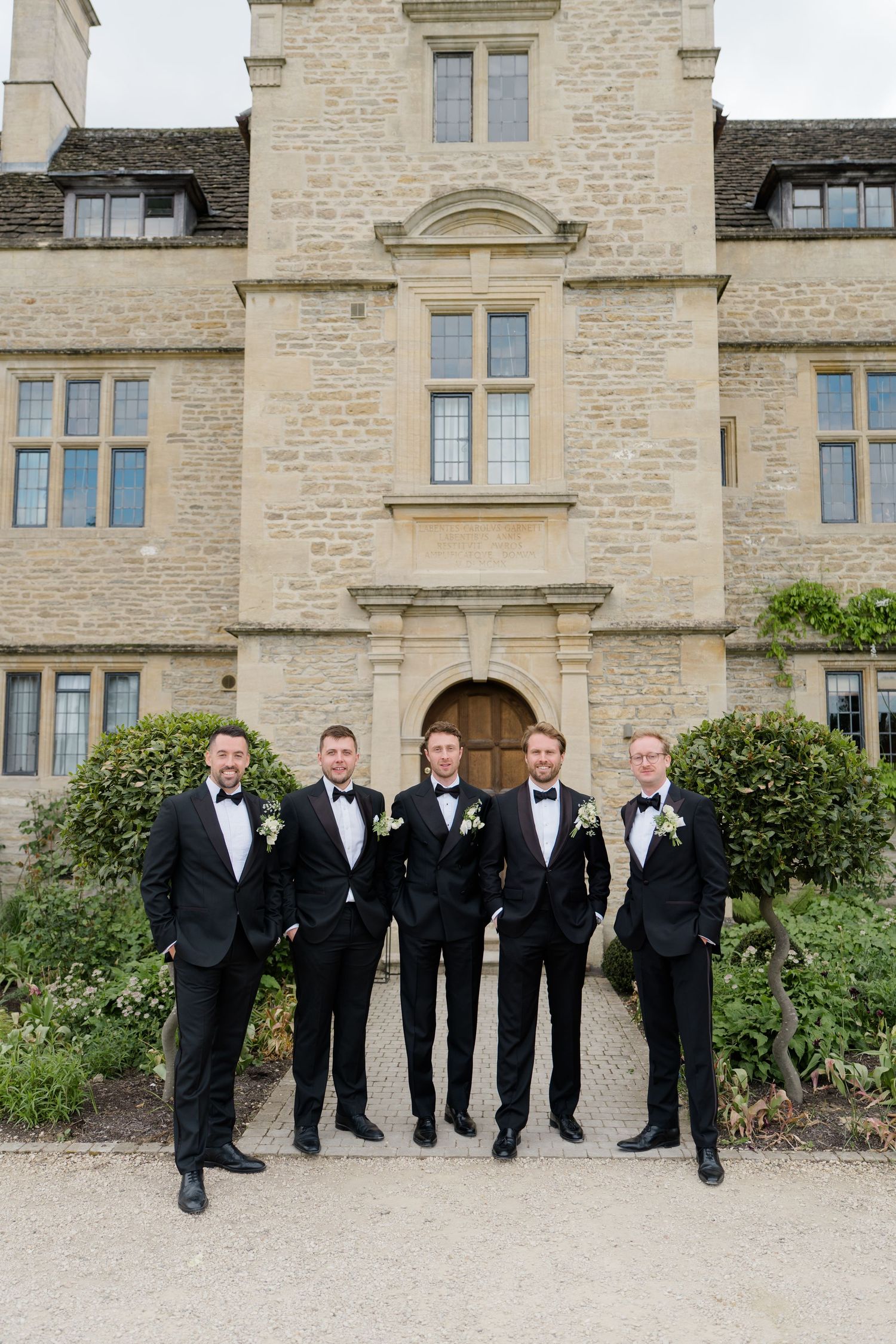 Five groomsmen in matching black tuxedos with bow ties and boutonnieres posing before a historic stone manor.