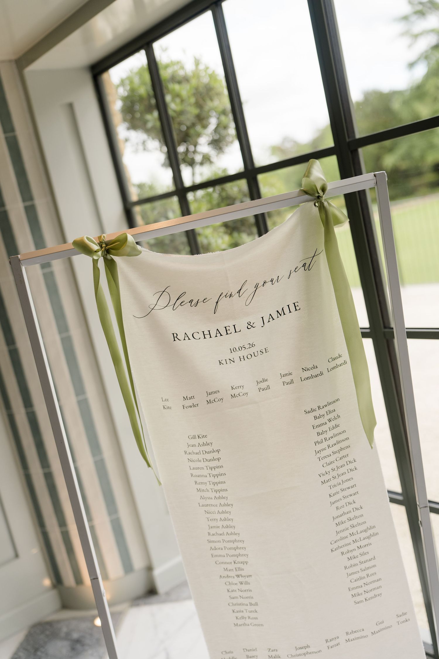 Elegant fabric wedding seating chart for Rachael and Jamie displayed on a stand with green ribbon bows.