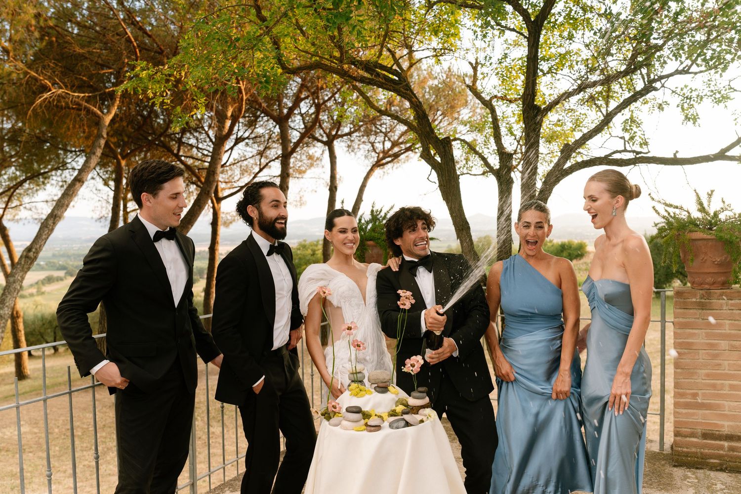 Wedding party gathered around a champagne table under trees at sunset.