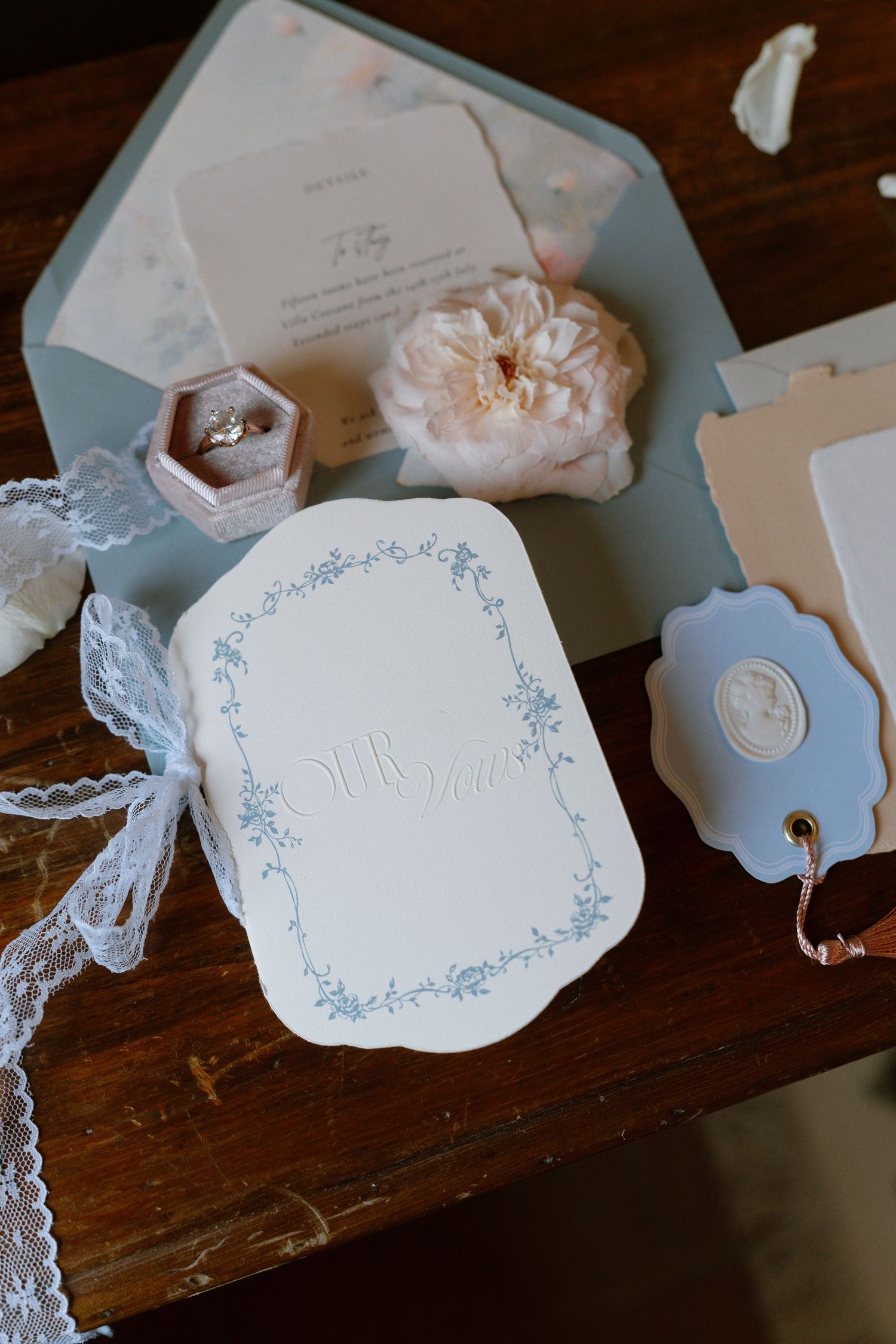 Delicate white wedding stationery with blue floral borders and lace details arranged on a dark wooden surface.