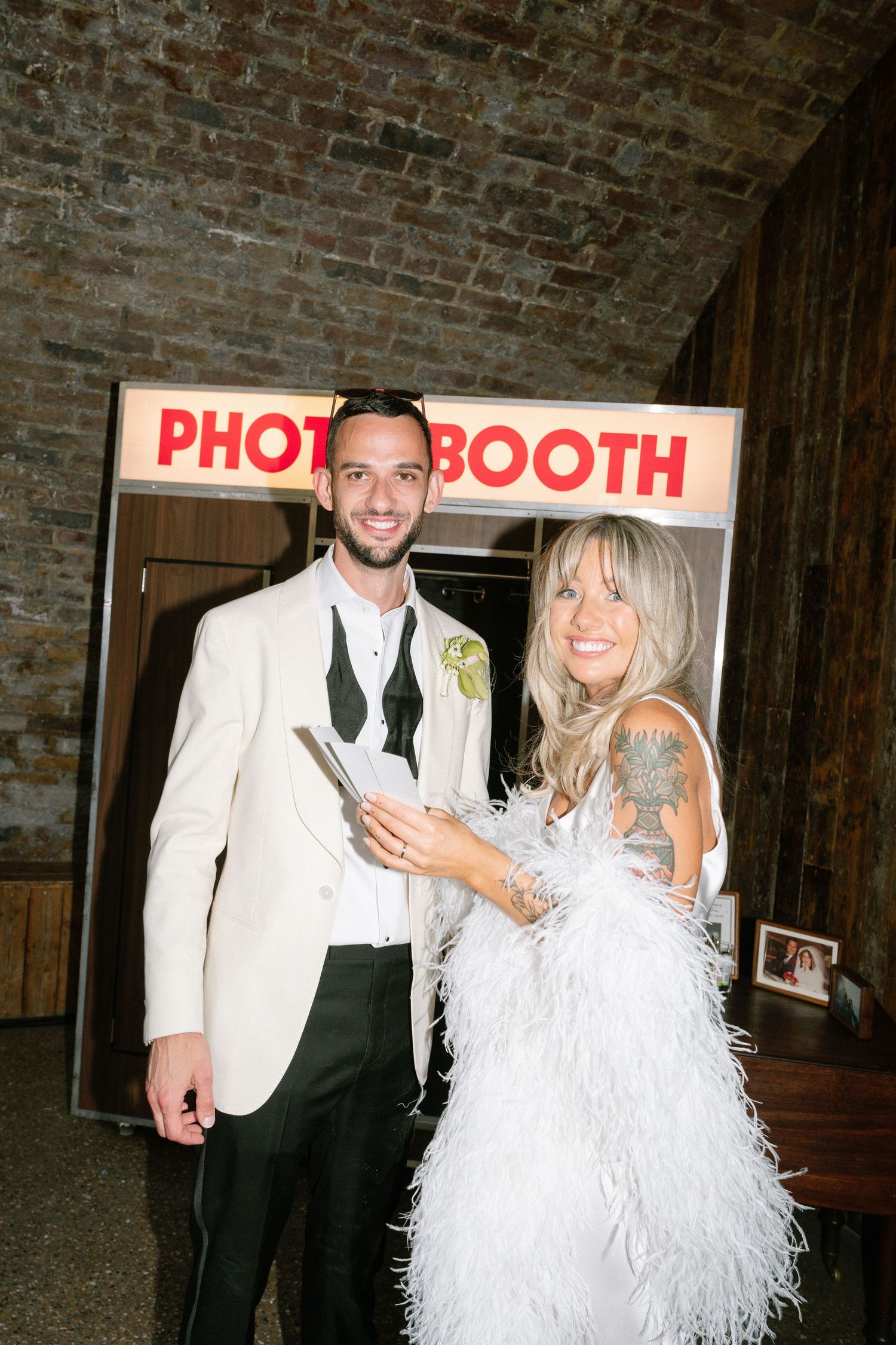 A couple poses in front of a photo booth at a wedding celebration with a white feather boa and formal attire.