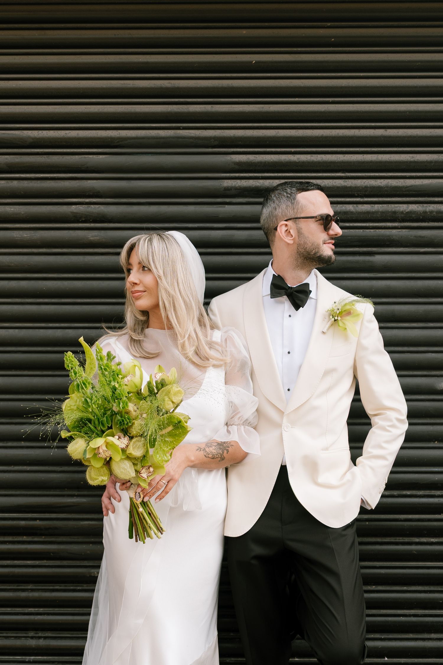 Wedding couple in white and black formal attire posing against an industrial metal backdrop.