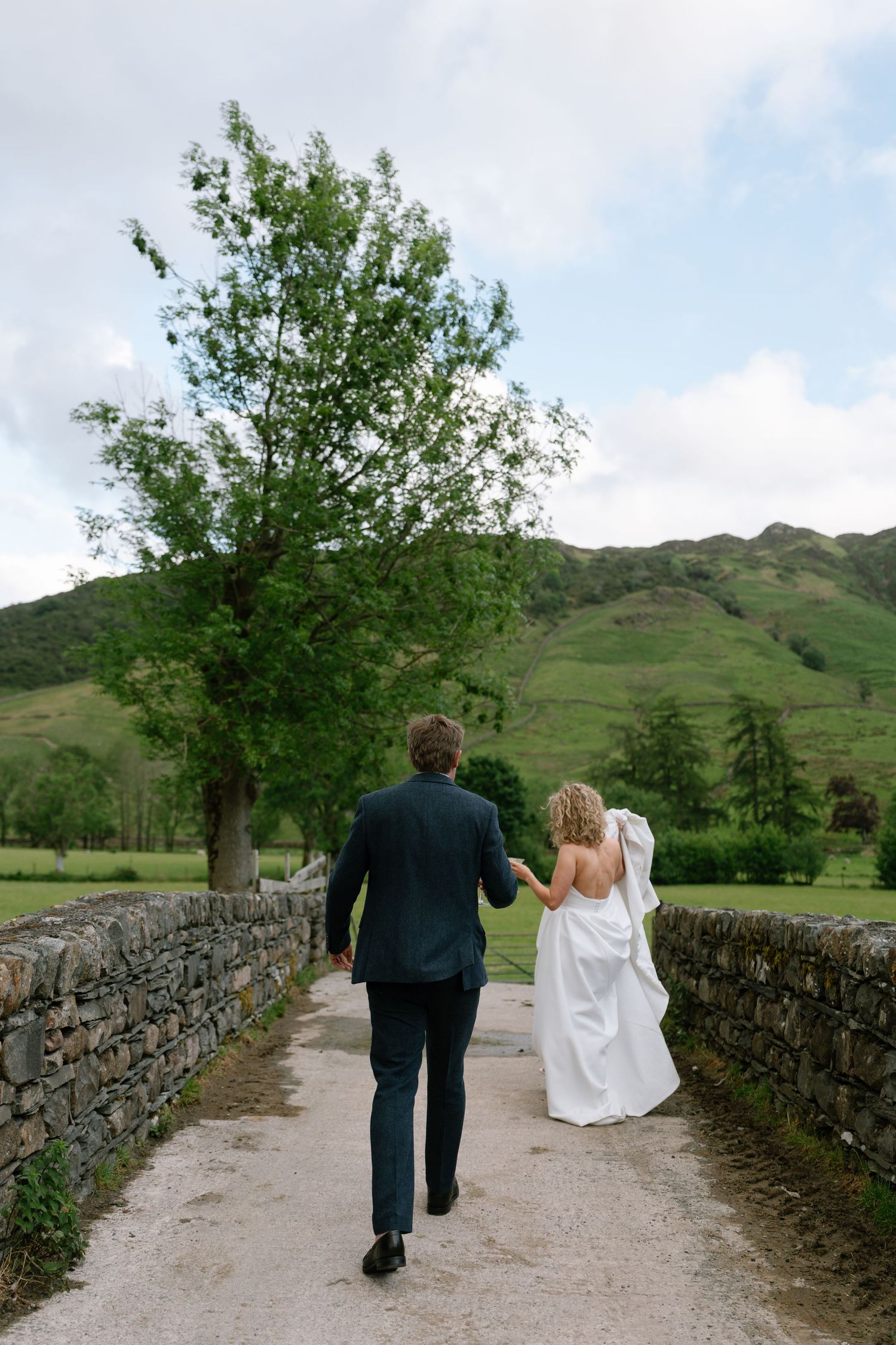 Newlyweds walk hand in hand along a stone-walled path with green hills in the background.