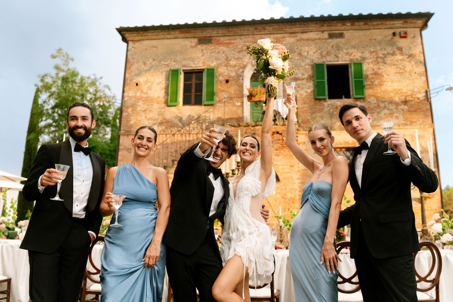 Wedding party celebrating and posing together outside a rustic Italian villa on a sunny day.