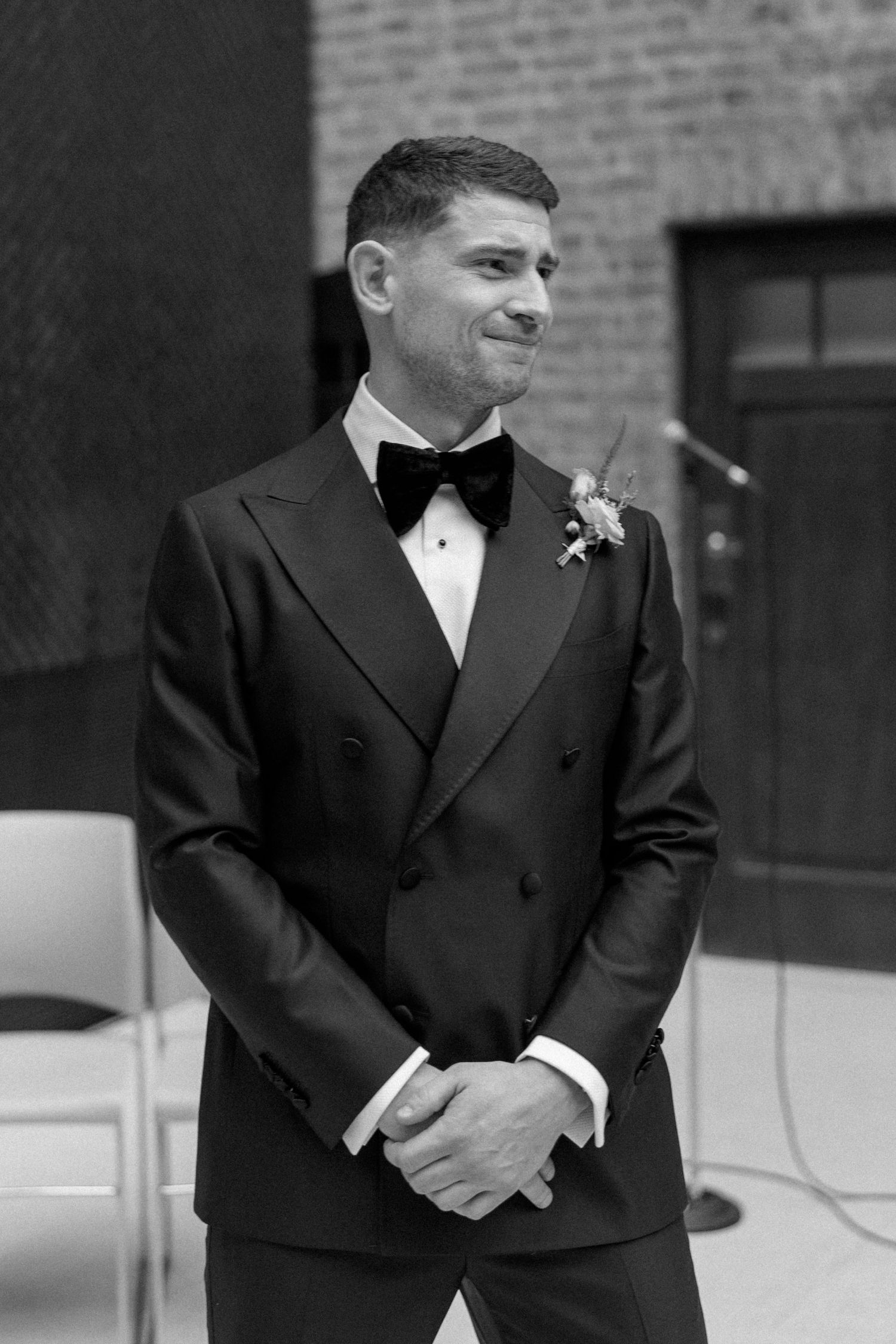 A groom in a black tuxedo and bowtie smiles while standing outdoors in a formal wedding photo.