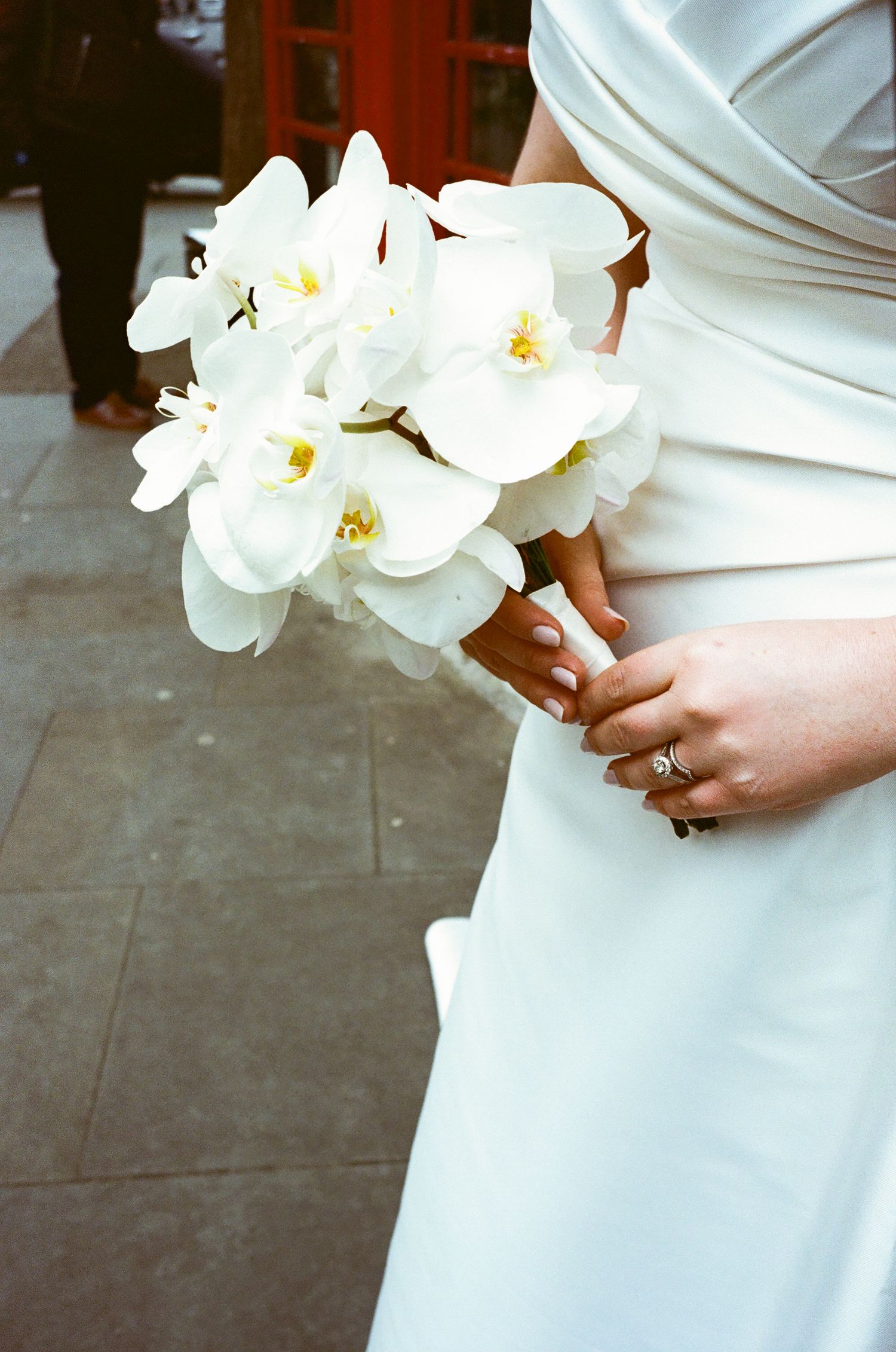 A white orchid bridal bouquet held against an elegant white wedding dress with a ring visible on the hand.