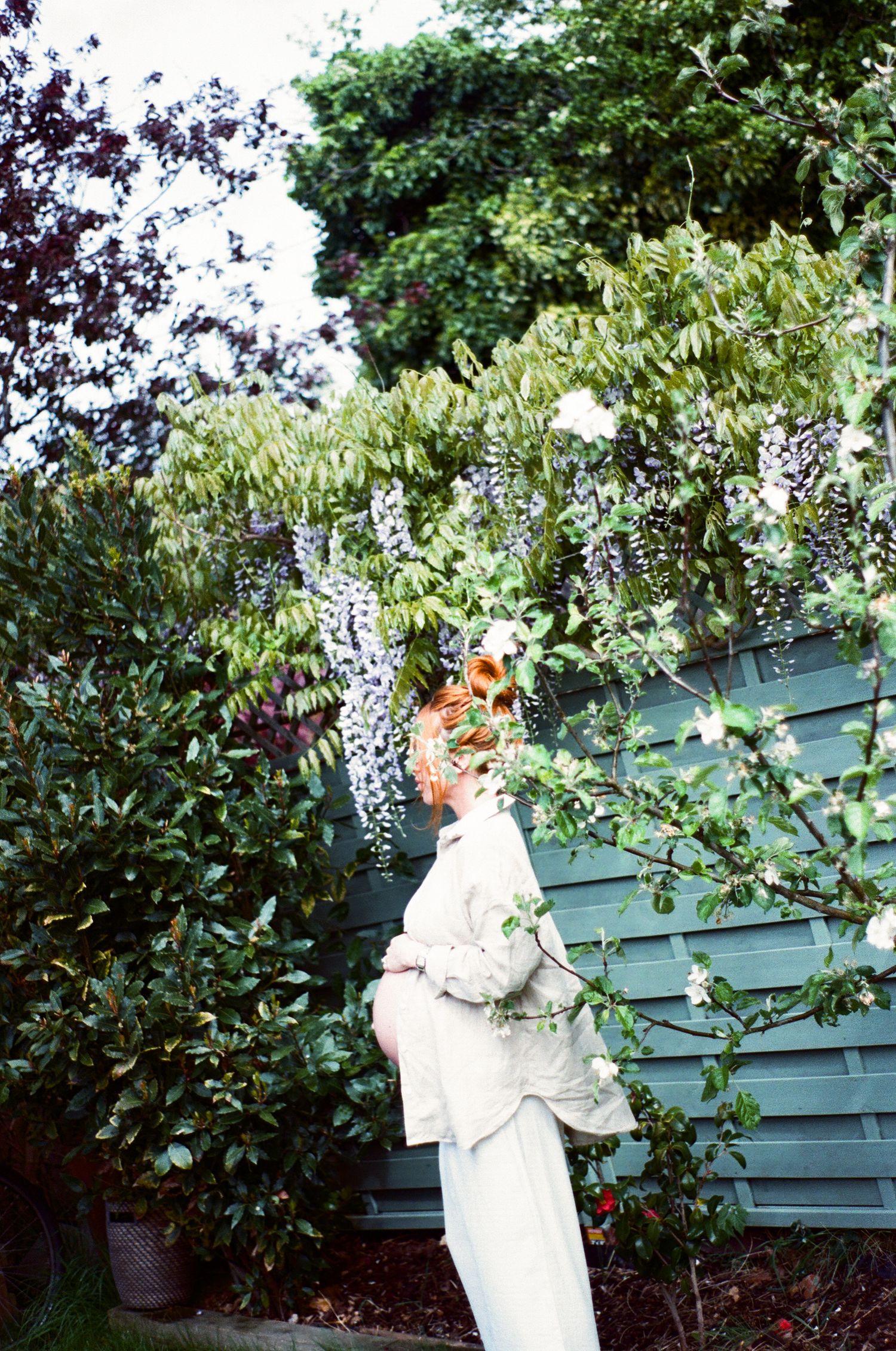 White statue holding flowers stands against a green garden fence with lush foliage in the background.