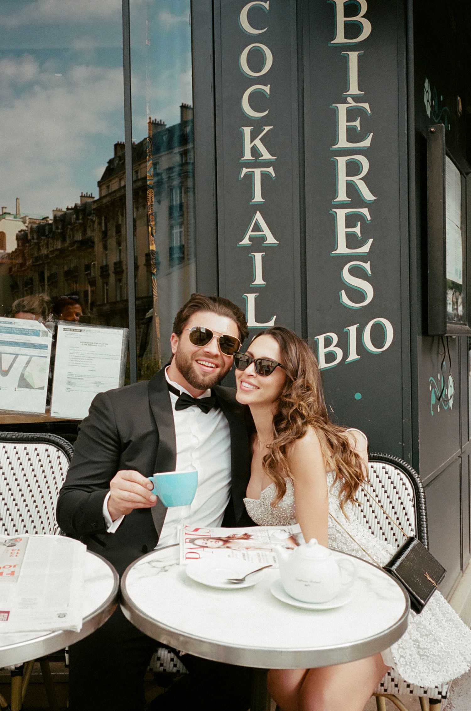A couple in formal attire enjoying coffee at a Parisian bistro with a vintage storefront sign reading 'Cocktail Bieres Bio'.