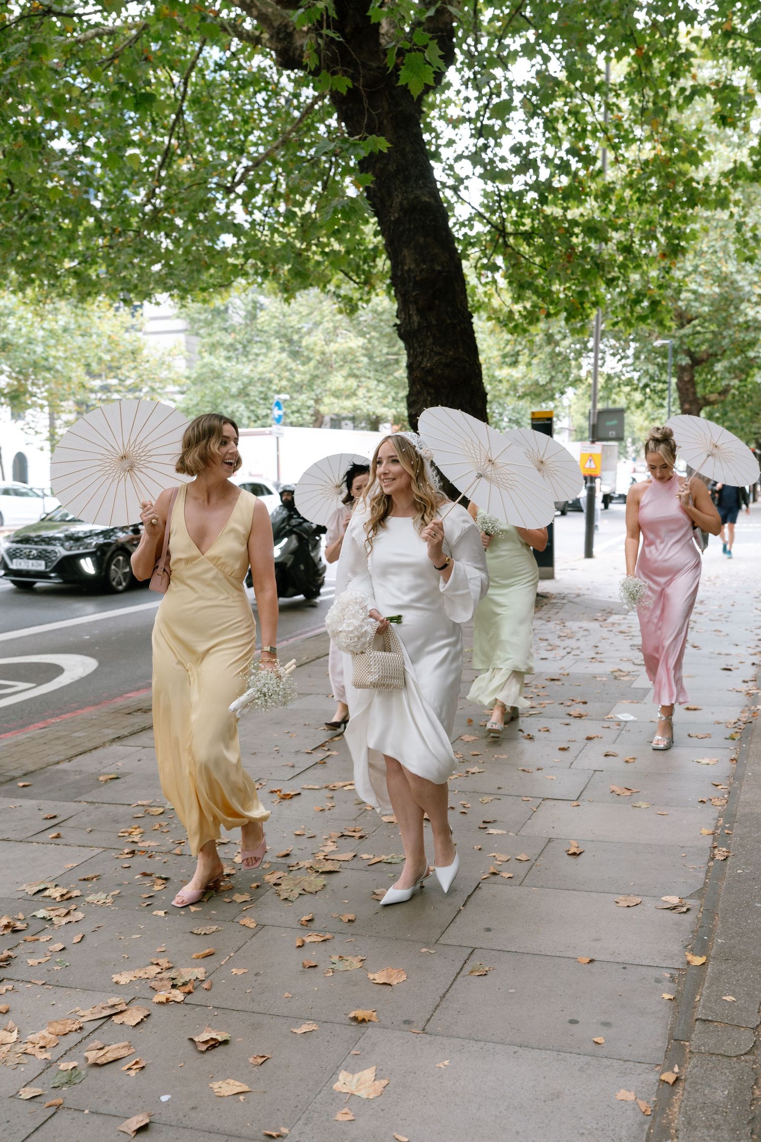 Wedding party walking along a tree-lined street holding white parasols on a sunny day.