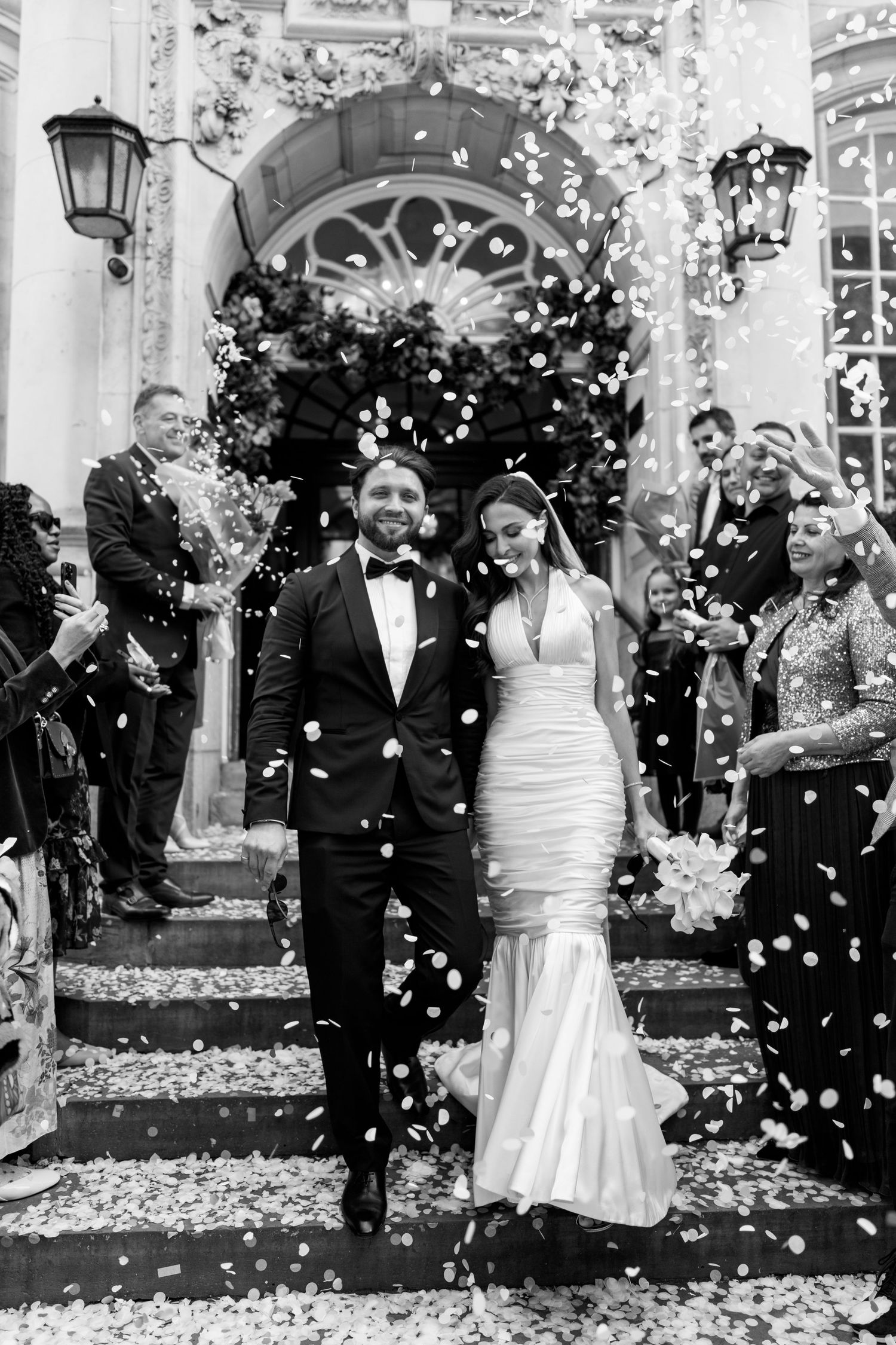 Wedding guests shower the newlyweds with white flower petals outside an elegant venue in a series of celebratory photos.