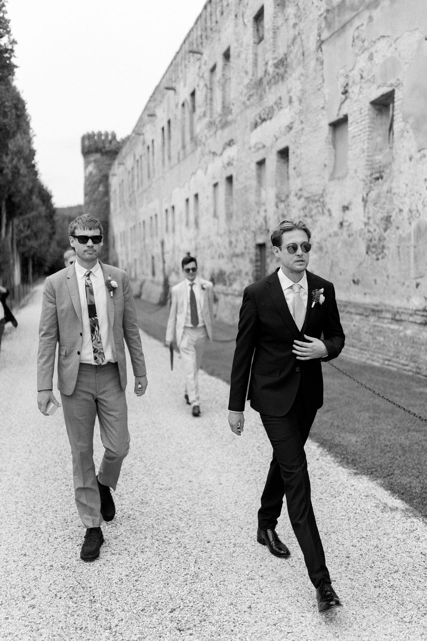 Groomsmen in suits walk along a historic stone wall pathway.