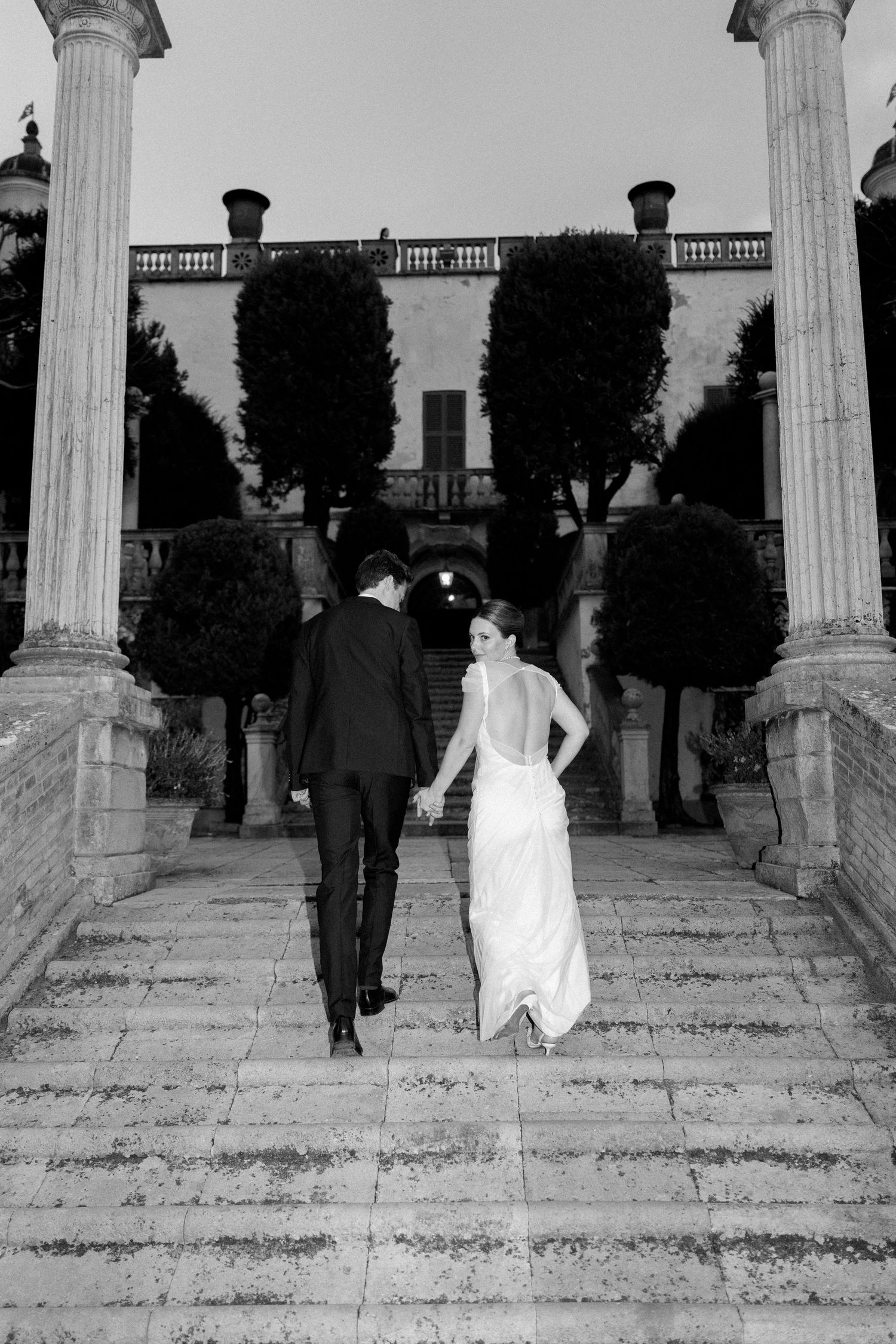 Wedding couple ascends stone steps between classical white columns at historic venue.