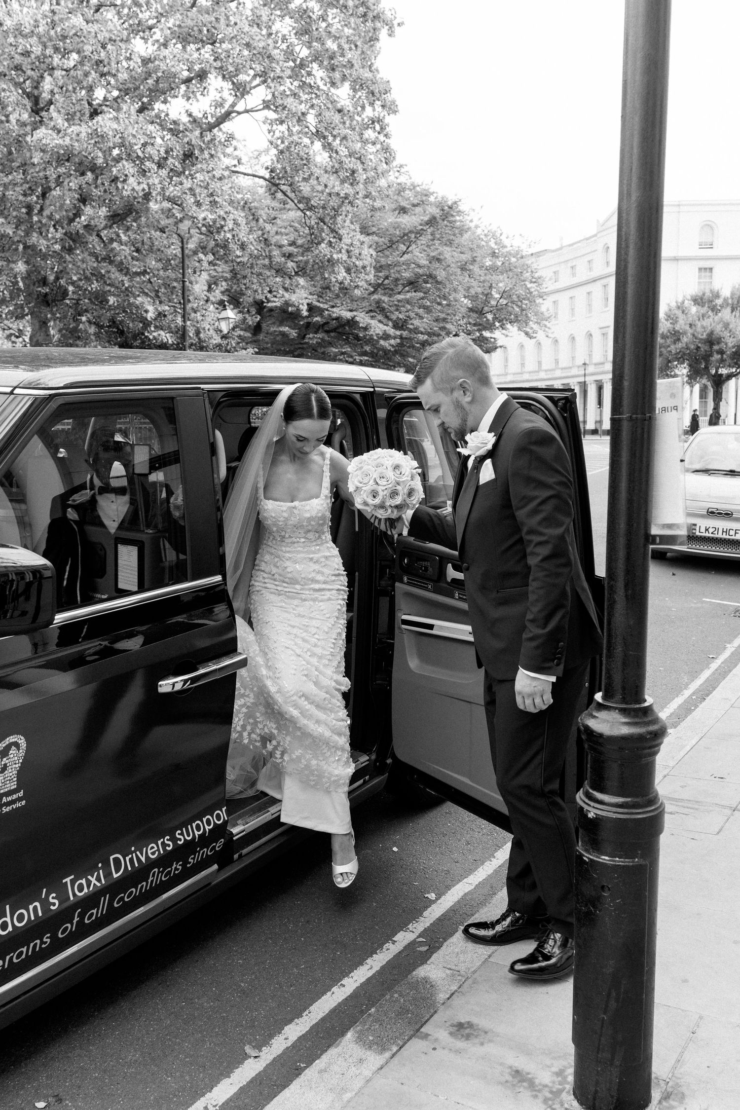 A wedding couple steps out of a London taxi cab with the bride carrying a white floral bouquet.