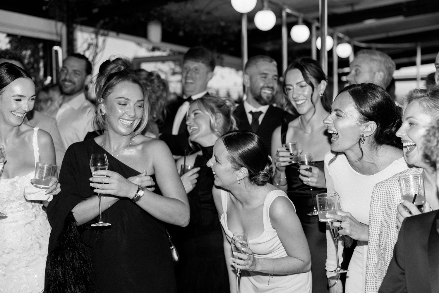 Wedding guests laugh and socialize while holding champagne glasses at an indoor reception celebration.