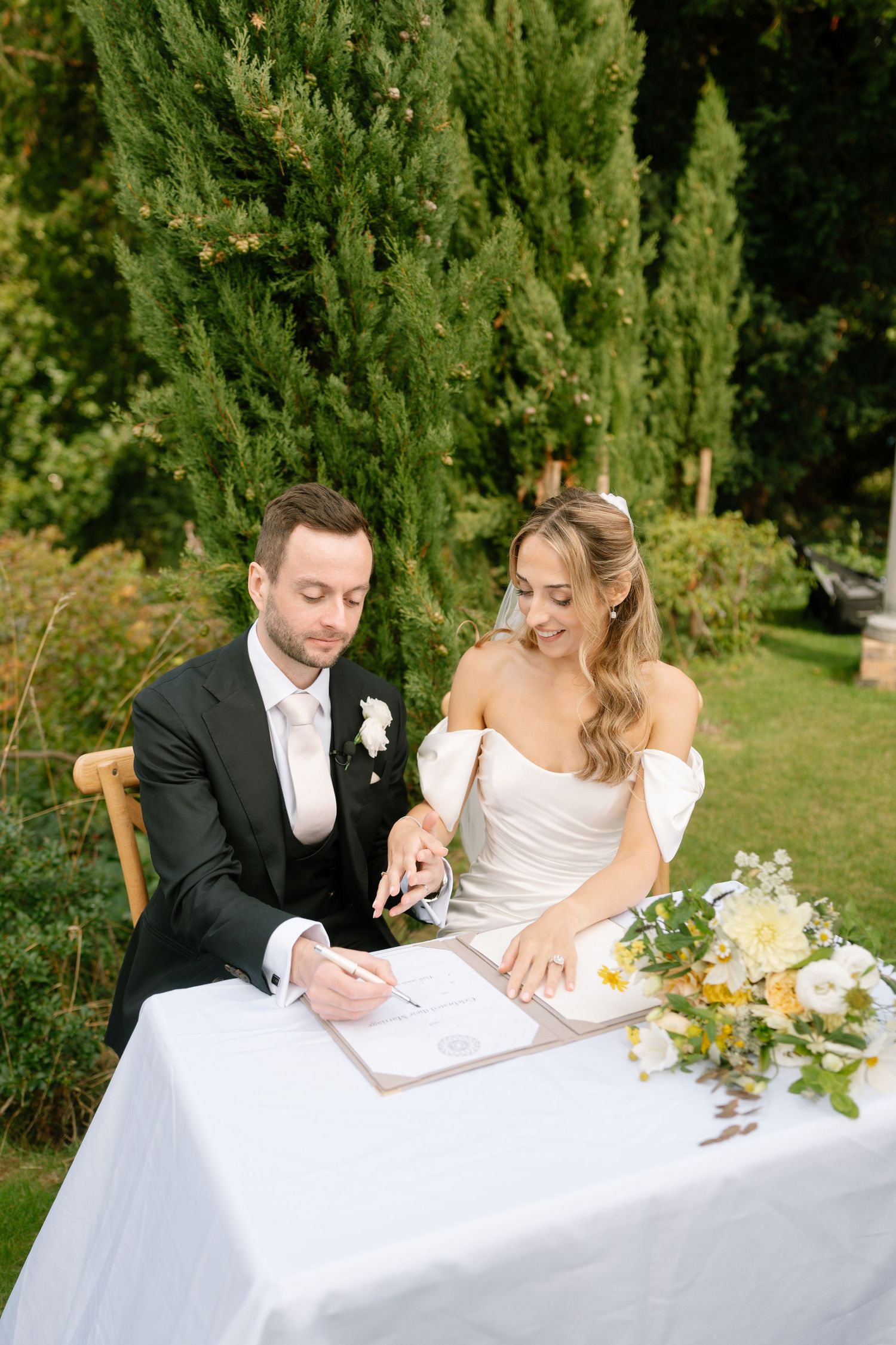 Couple signing marriage documents at a white-clothed table outdoors with greenery in background.