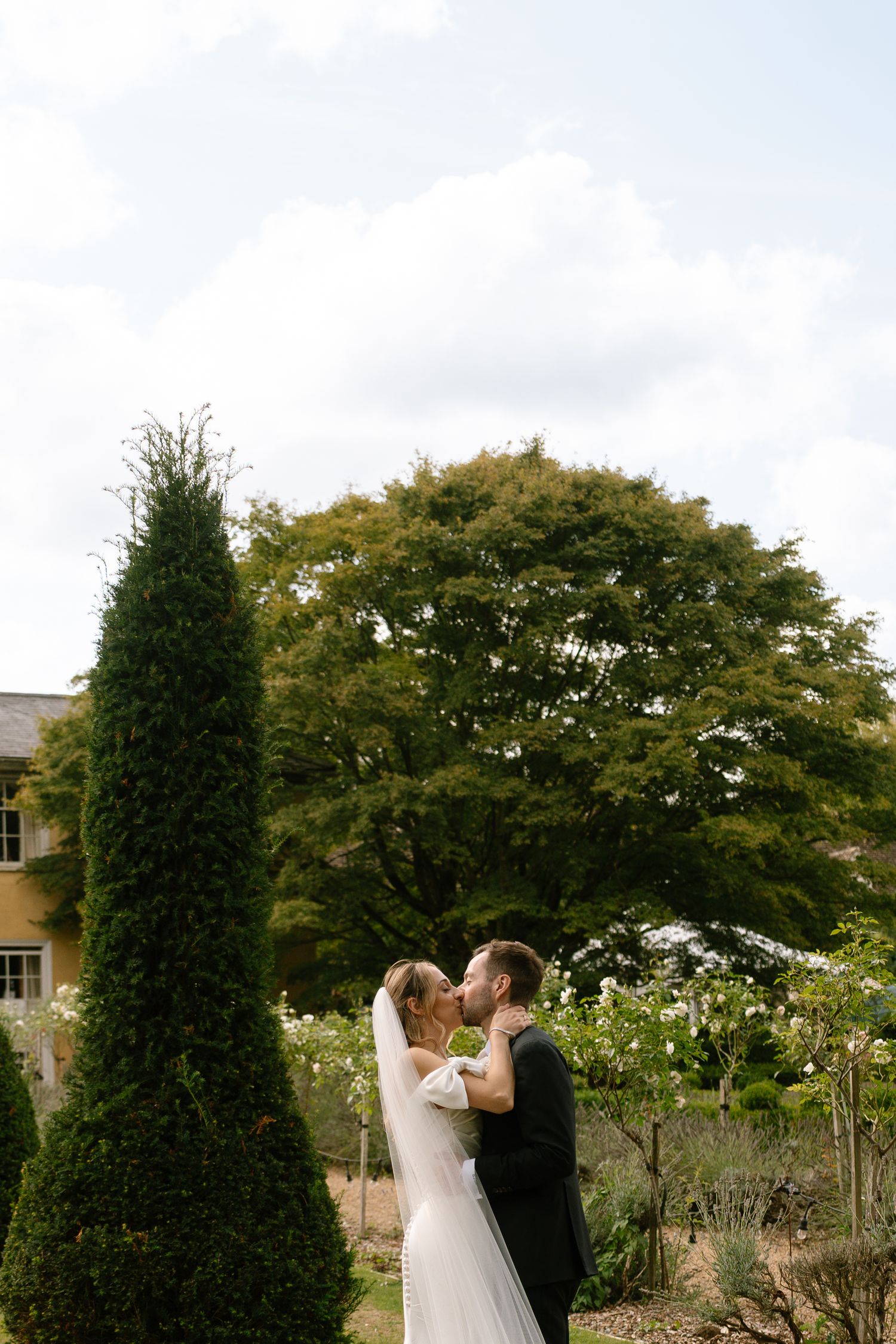 A romantic wedding photoshoot in a garden with tall evergreen trees and a yellow building in the background.