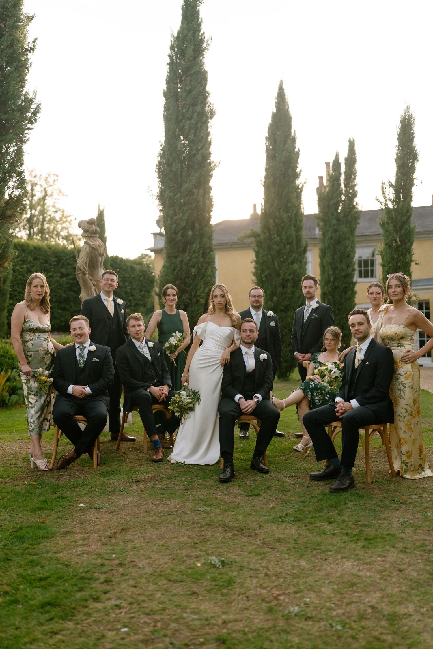 Wedding party poses together outdoors in front of tall cypress trees on a grassy lawn.