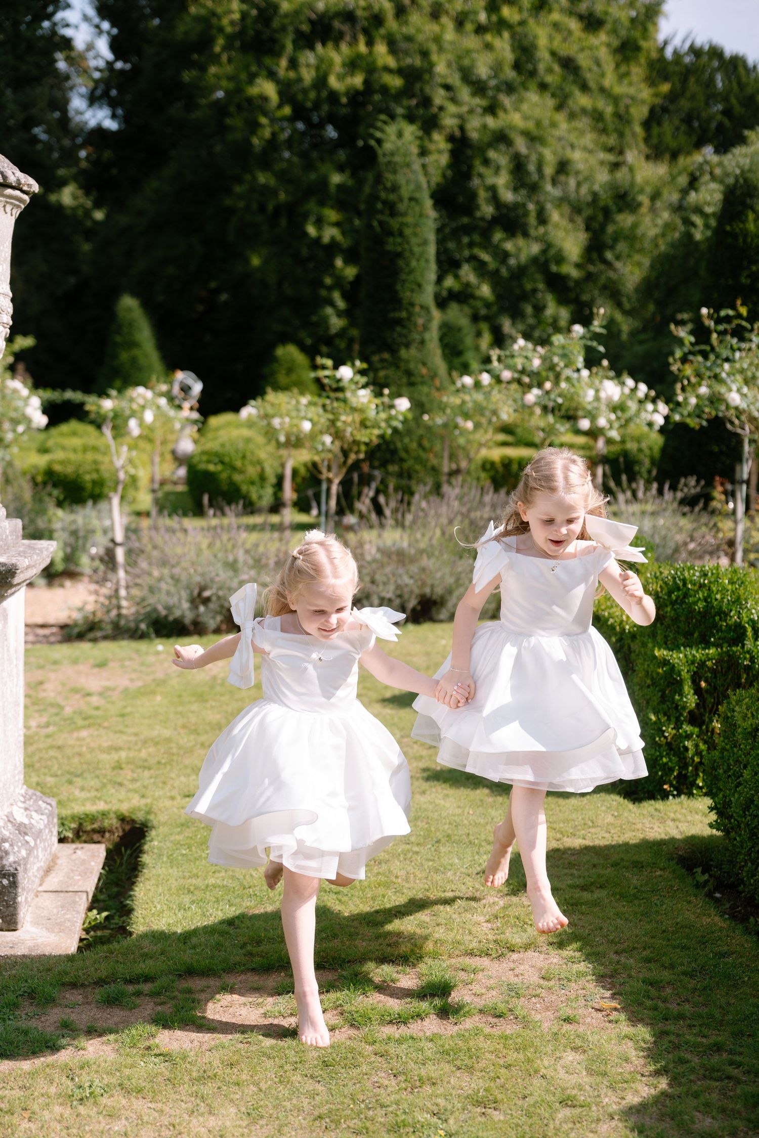 Young children in white dresses dance and twirl joyfully in a sunny garden setting.