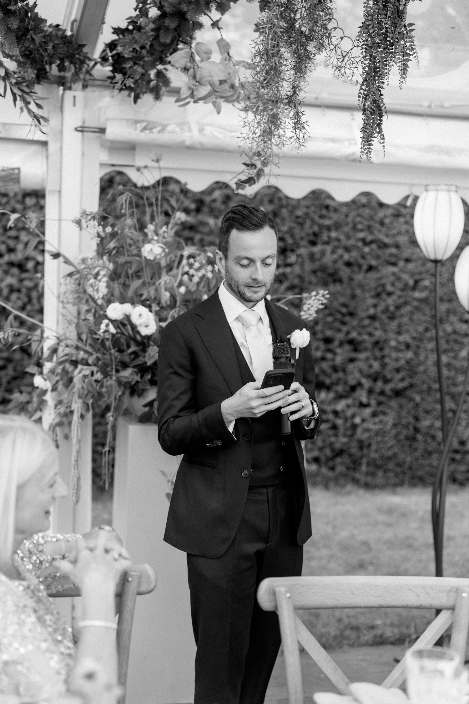 Wedding party member in formal attire checking phone under decorated tent canopy.