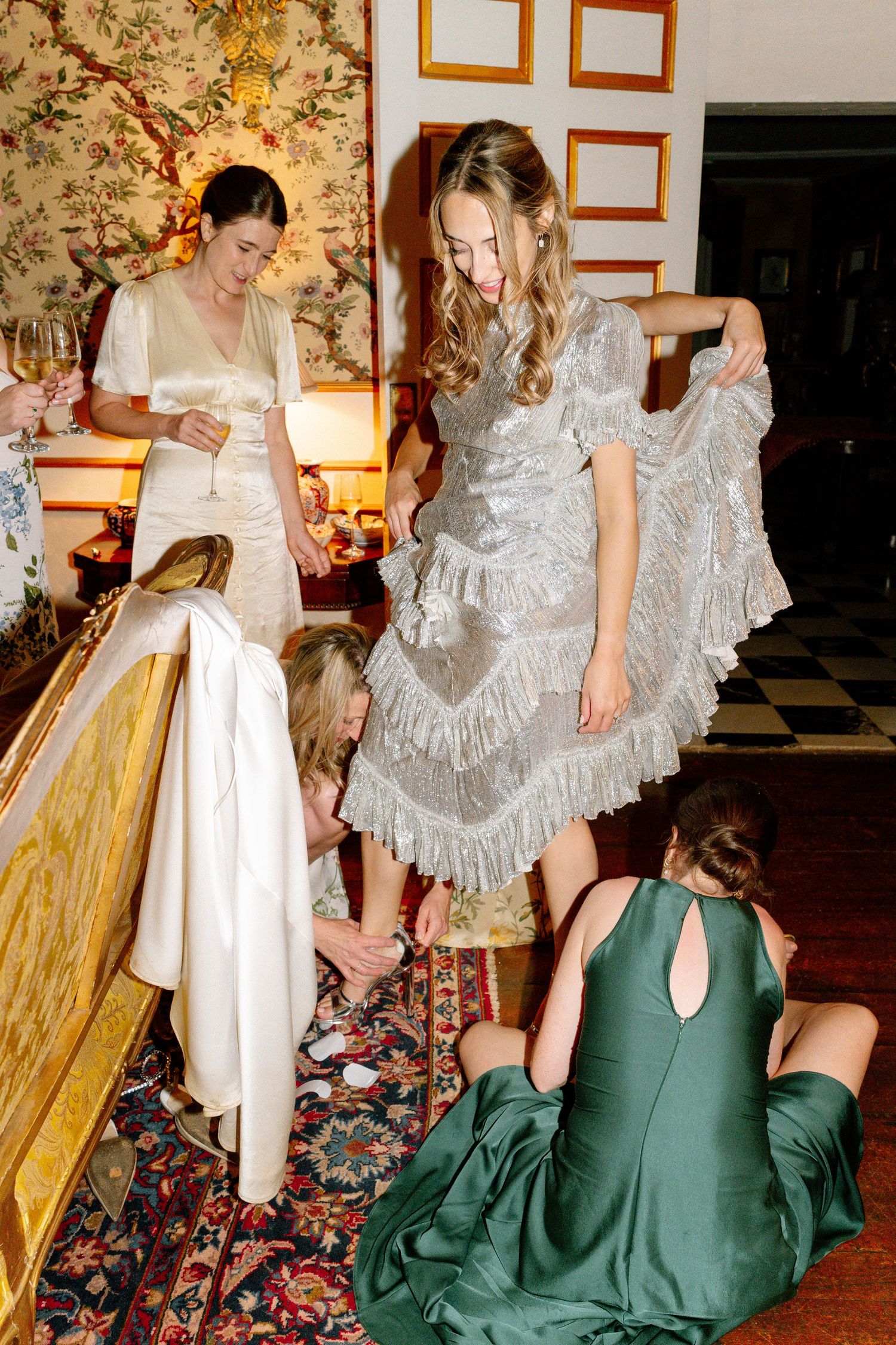 People in vintage-style dresses and gowns interact in an ornately decorated room with floral wallpaper.