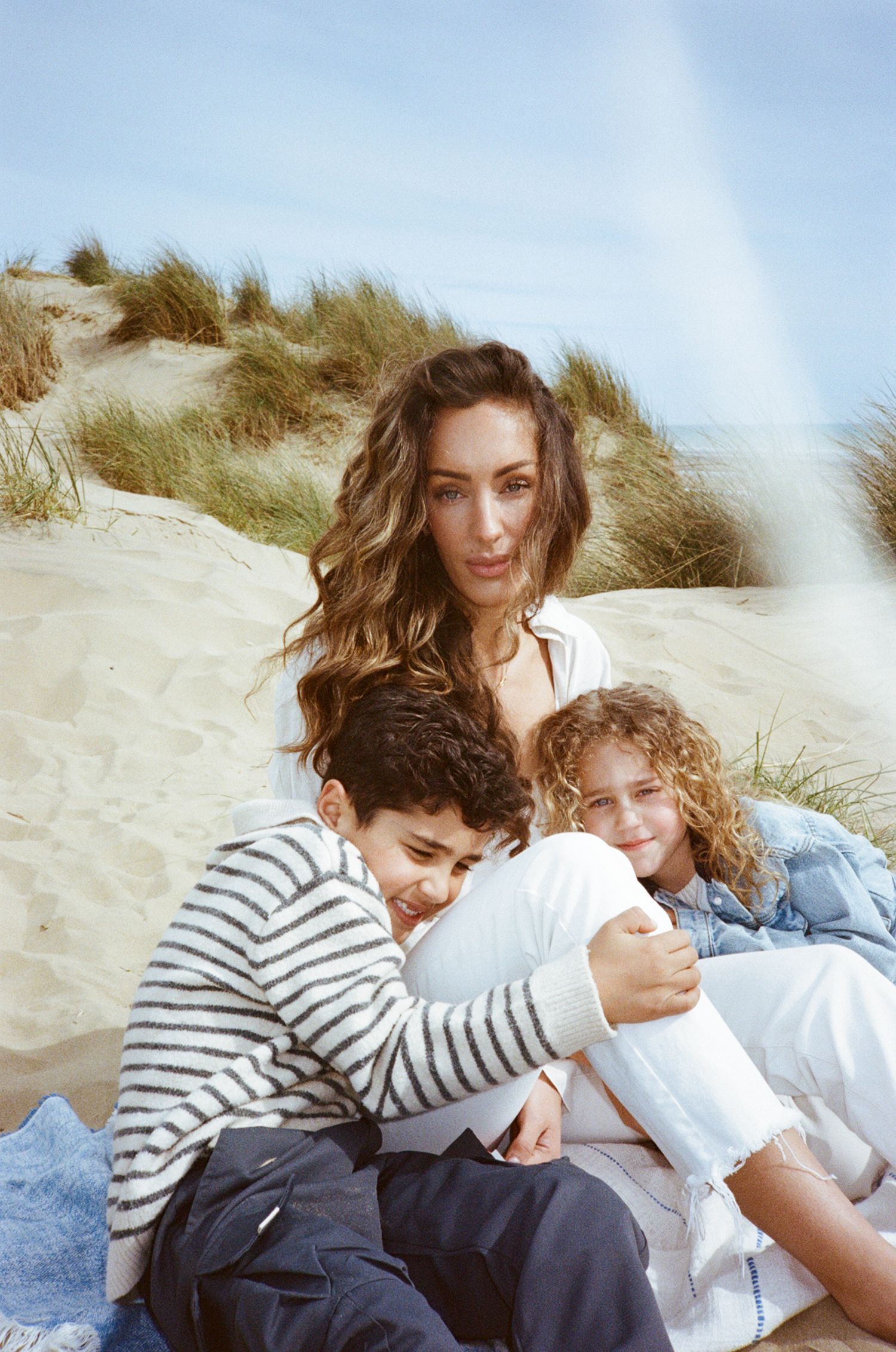 A woman with long wavy hair sits with two children on sandy beach dunes, sharing a warm embrace on a sunny day.
