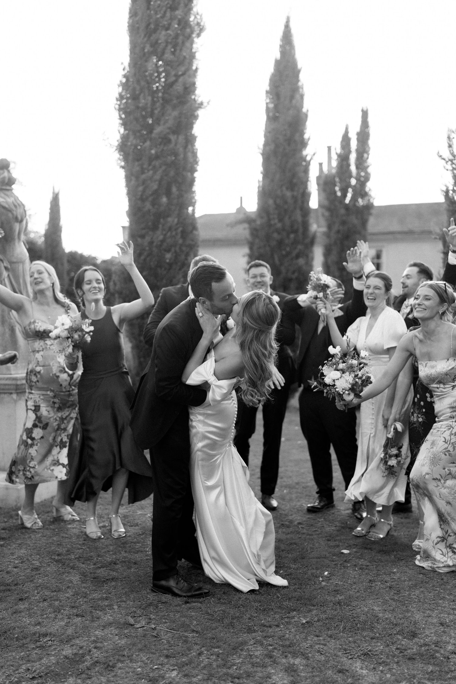 Wedding party dancing and celebrating on a lawn with cypress trees in the background.