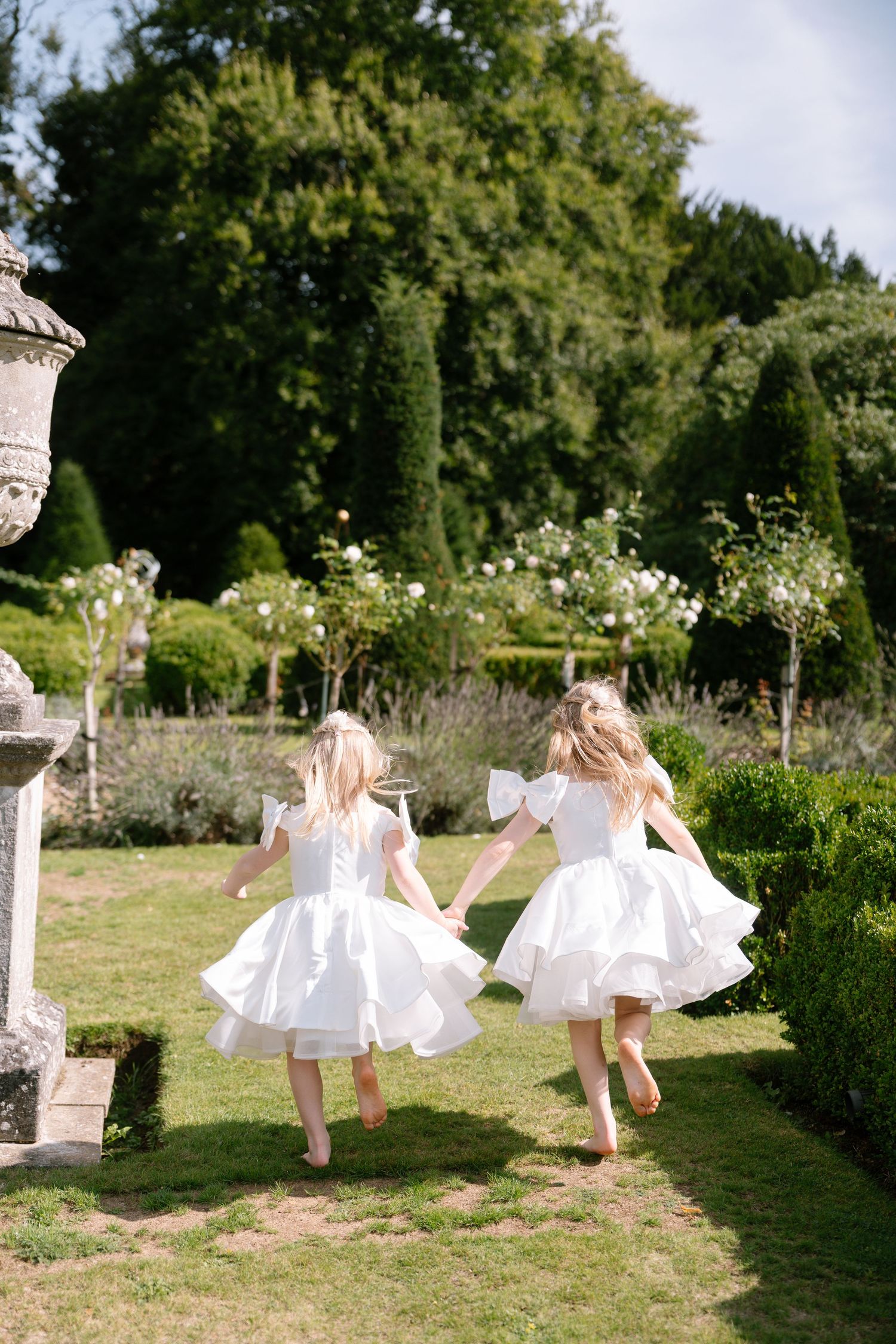 Children in matching white dresses run and play in a formal garden with stone pillars.