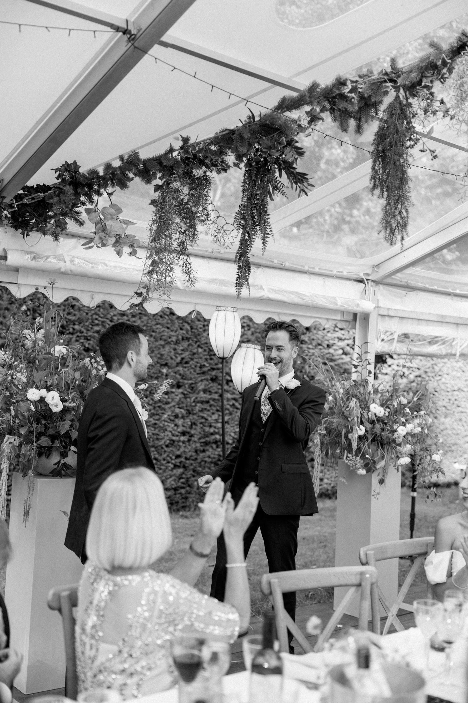 Wedding guests enjoying speeches during reception under greenery-decorated tent.