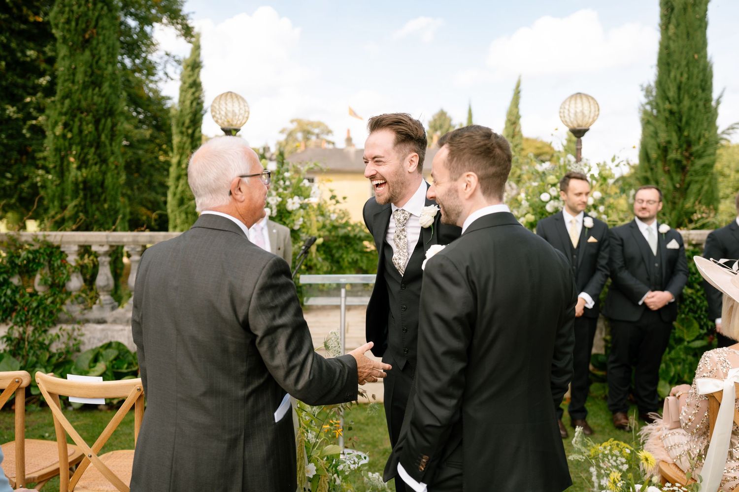 Groomsmen in black suits chatting before outdoor wedding ceremony.