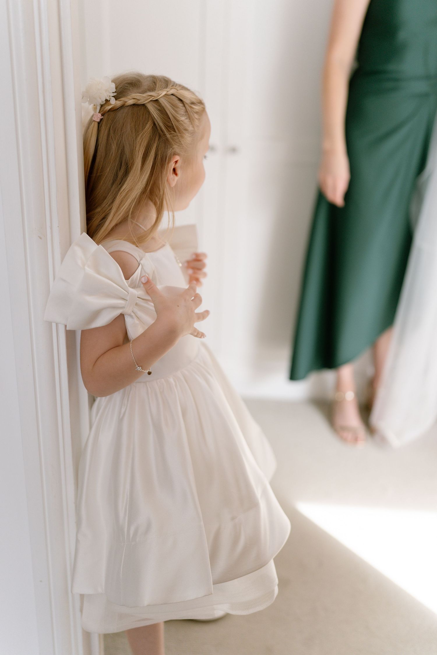 Young flower girl in white dress stands against wall near someone in green.