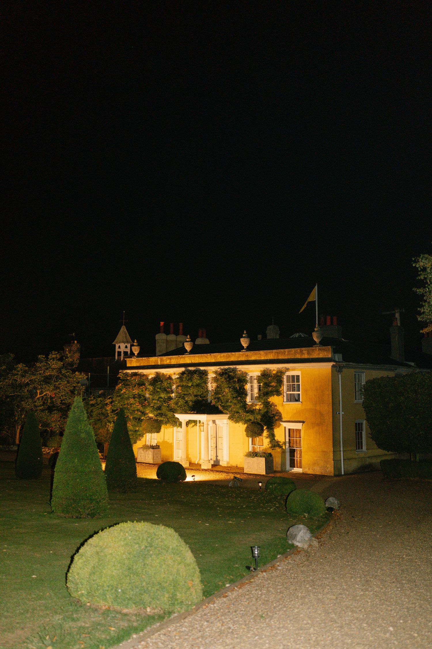 Illuminated yellow mansion with manicured gardens at night.