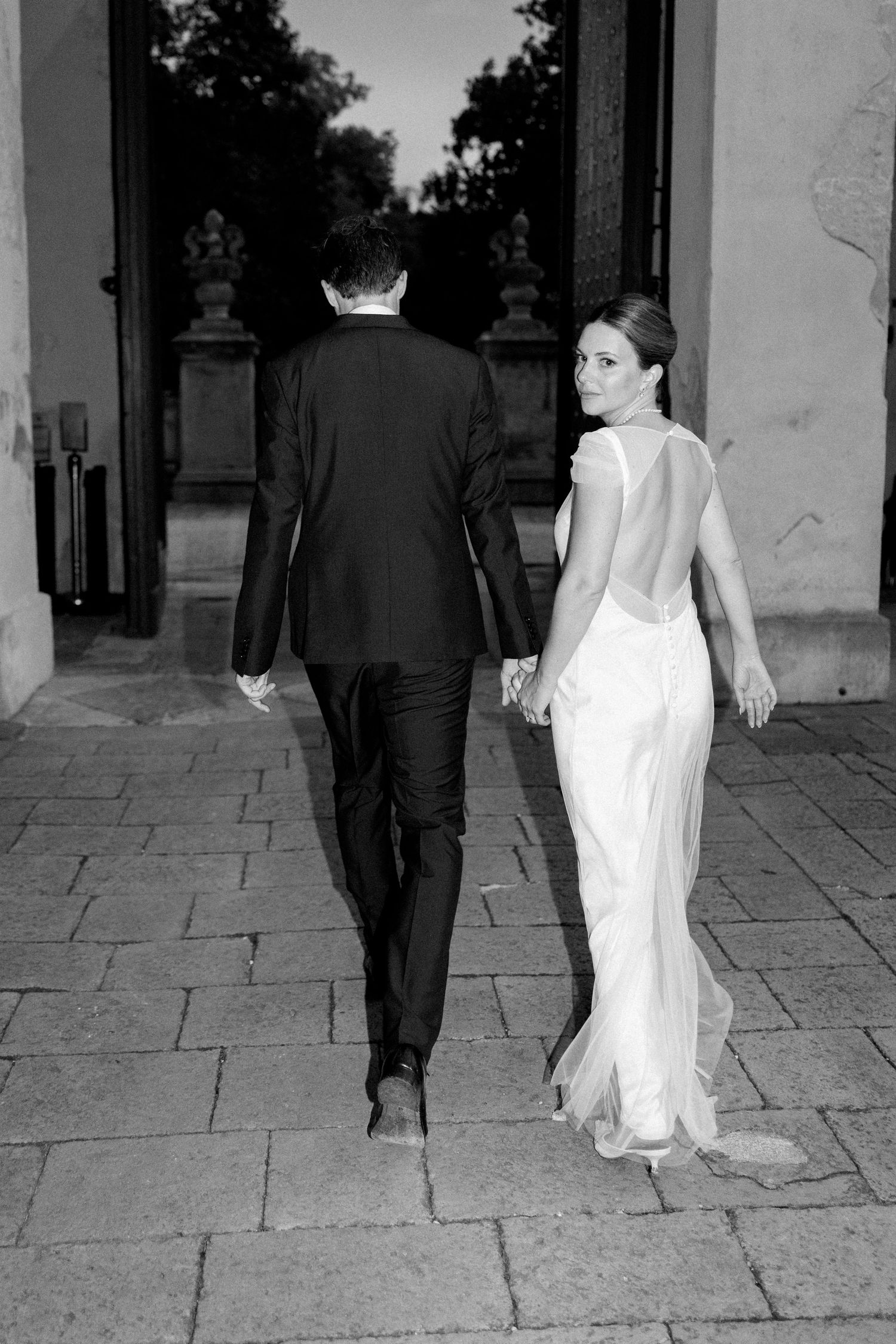 Bride in white gown and groom walk along stone pathway at elegant venue.