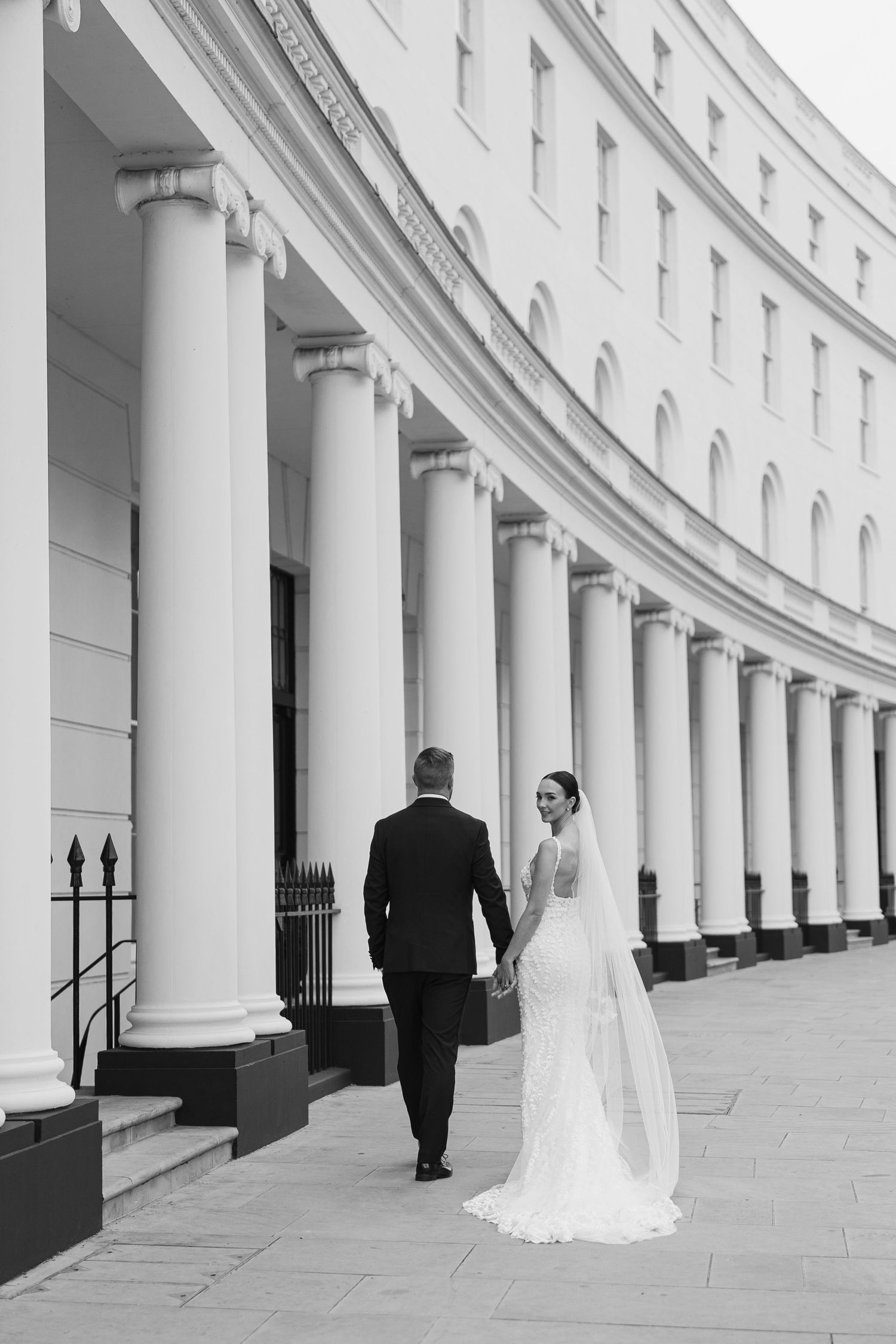 A bride and groom walk along a curved Georgian building with white columns in London.