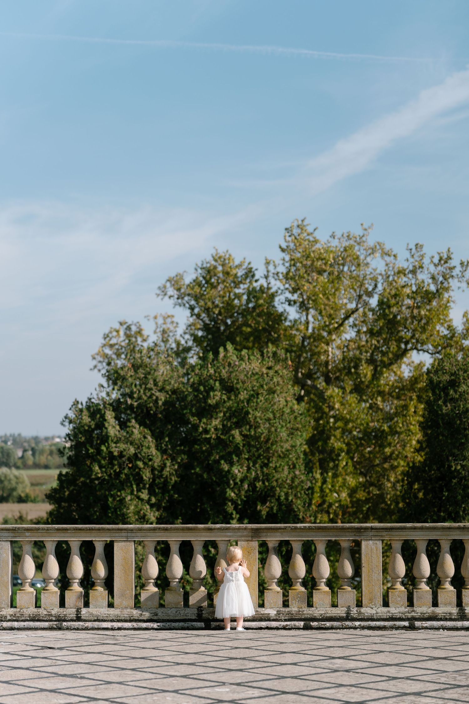 Small child in white dress stands alone on castle terrace with trees in background.