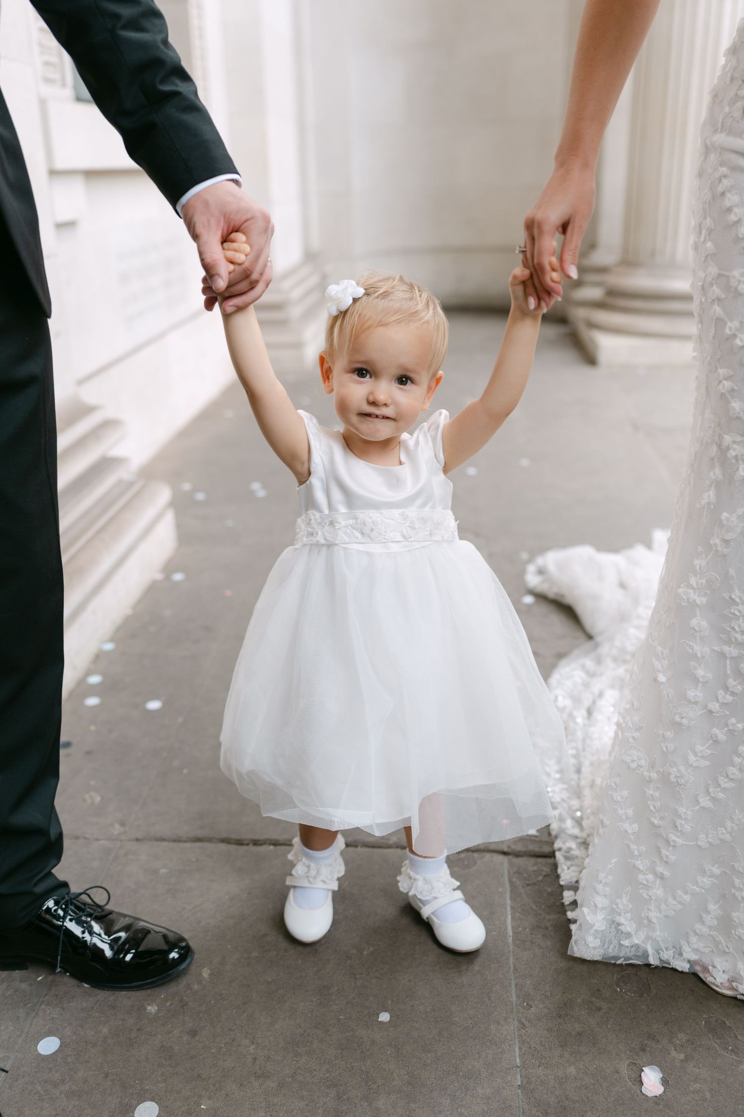 A small child in white dress holds hands with adults during a wedding.