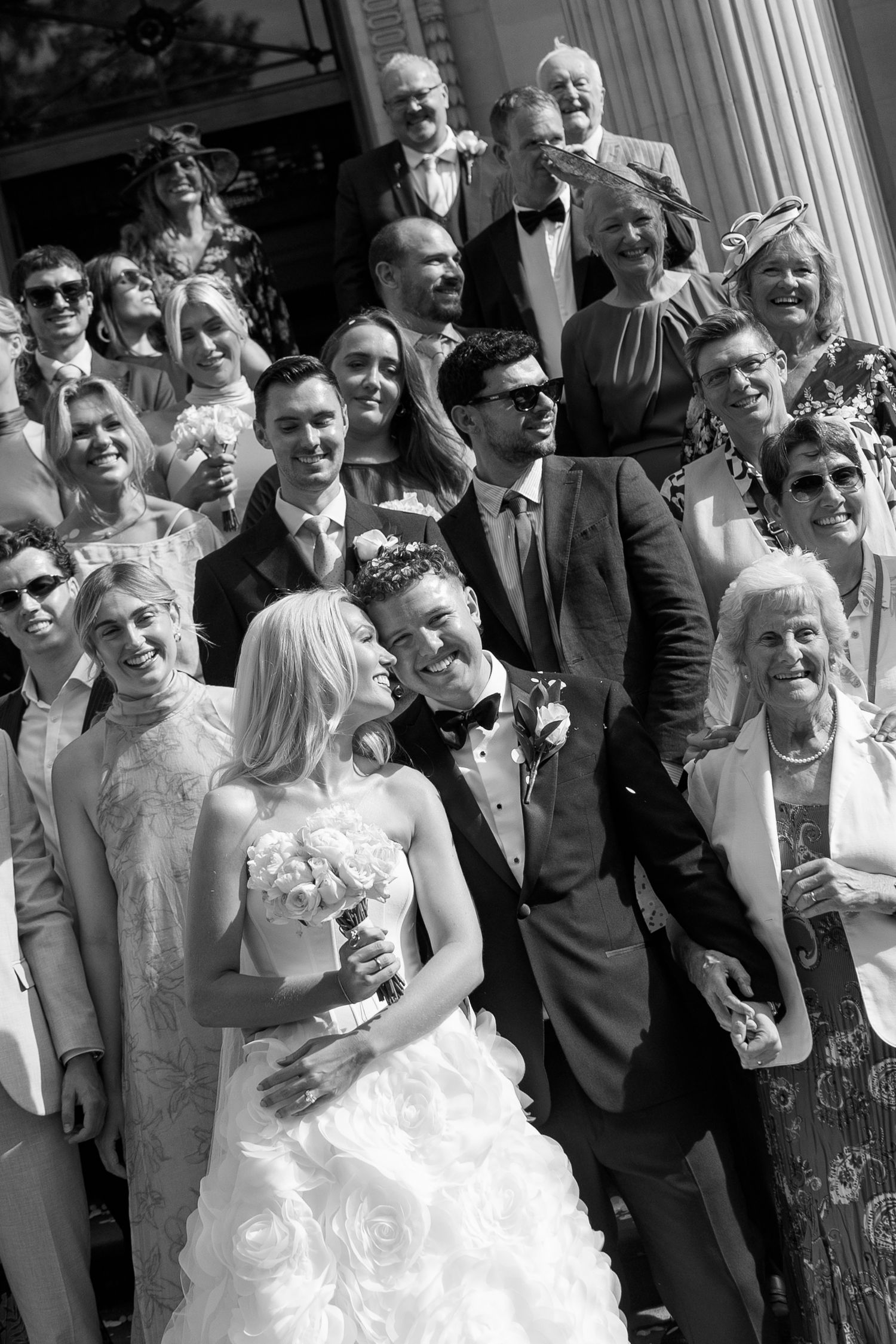 Black and white wedding photo of bride and groom kissing surrounded by smiling guests on church steps.