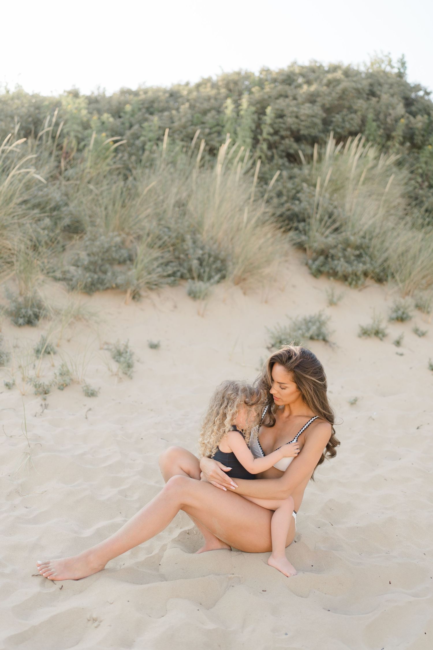 Intimate beach portrait of two people sitting together on sand dunes surrounded by coastal vegetation.