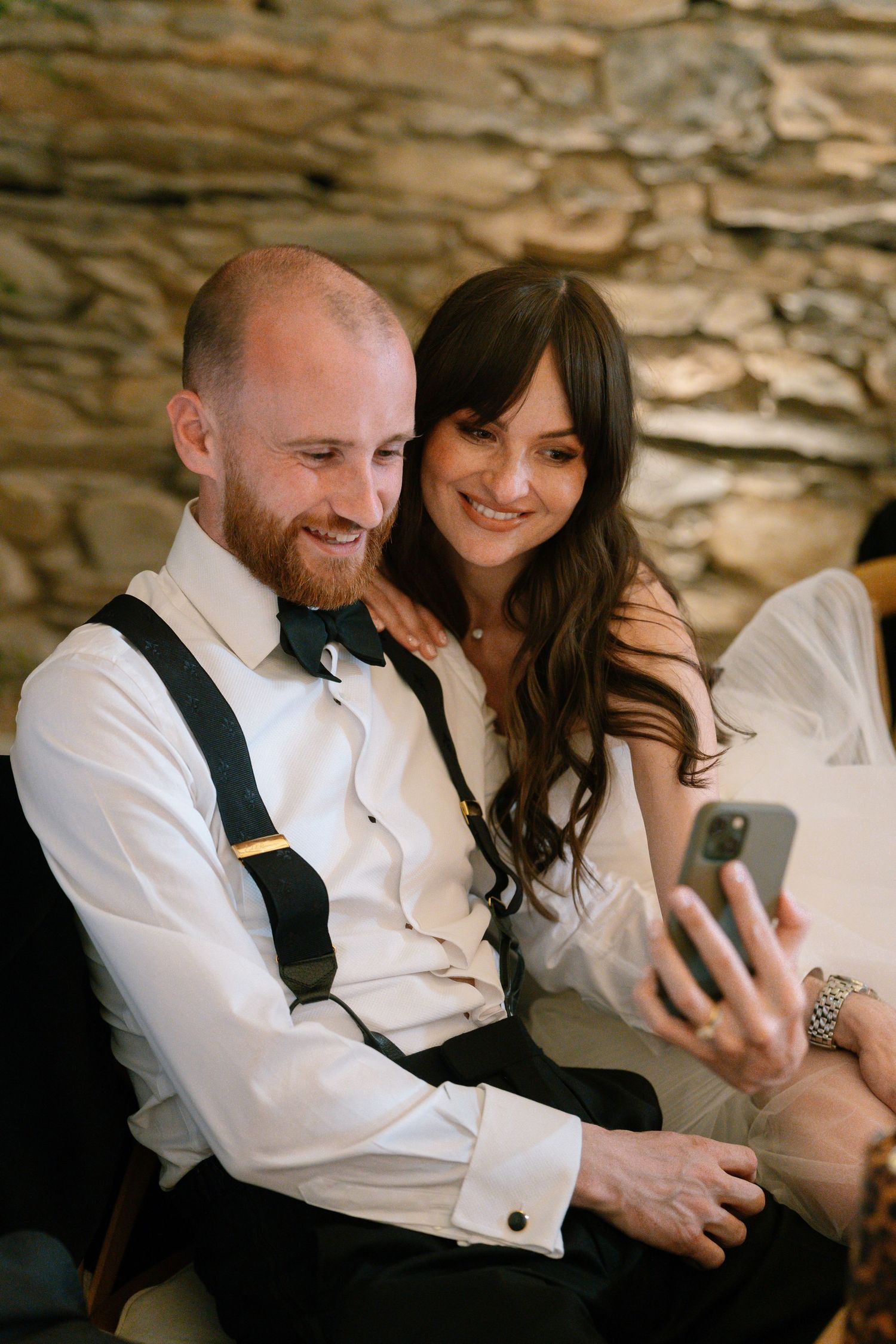 Wedding guests looking at a phone together while seated against a stone wall.
