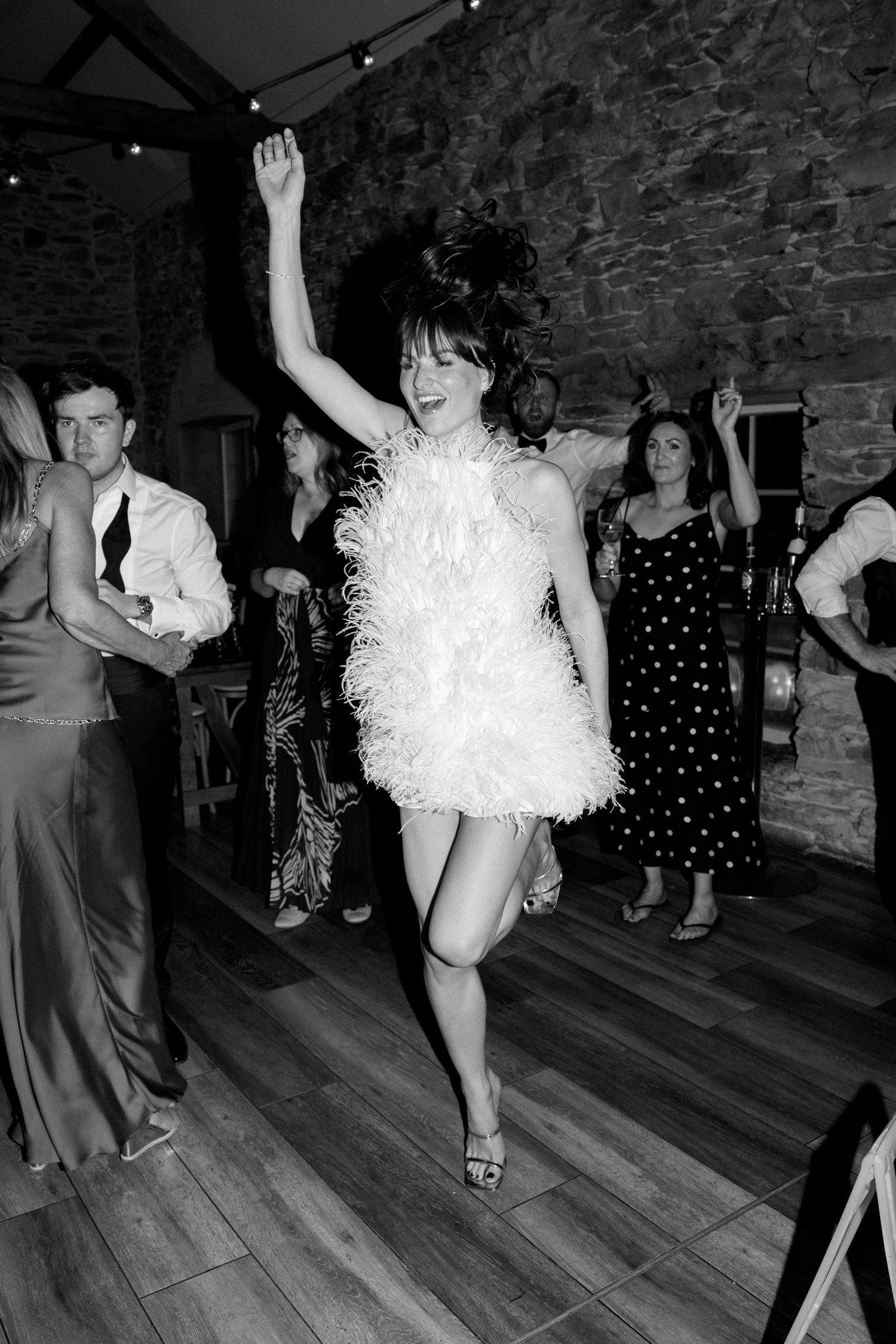 Dancers performing energetic moves on a wooden floor in a stone-walled venue with string lights.