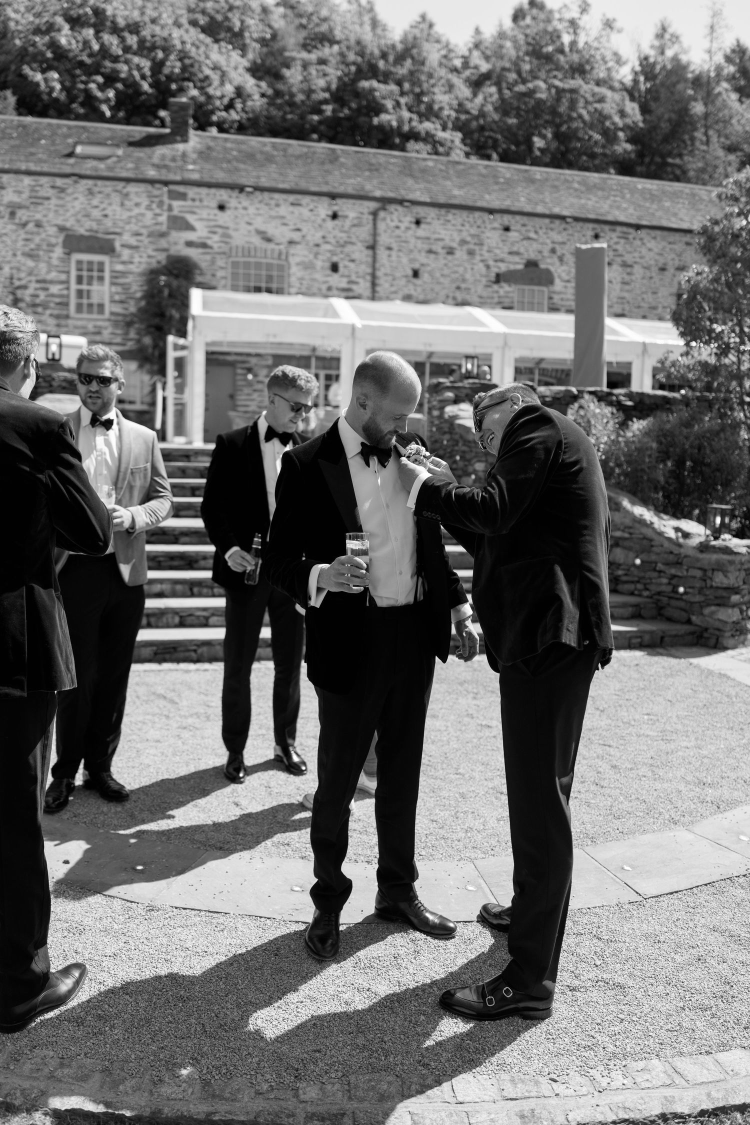 Groomsmen in tuxedos gather outside a stone building during a wedding celebration.