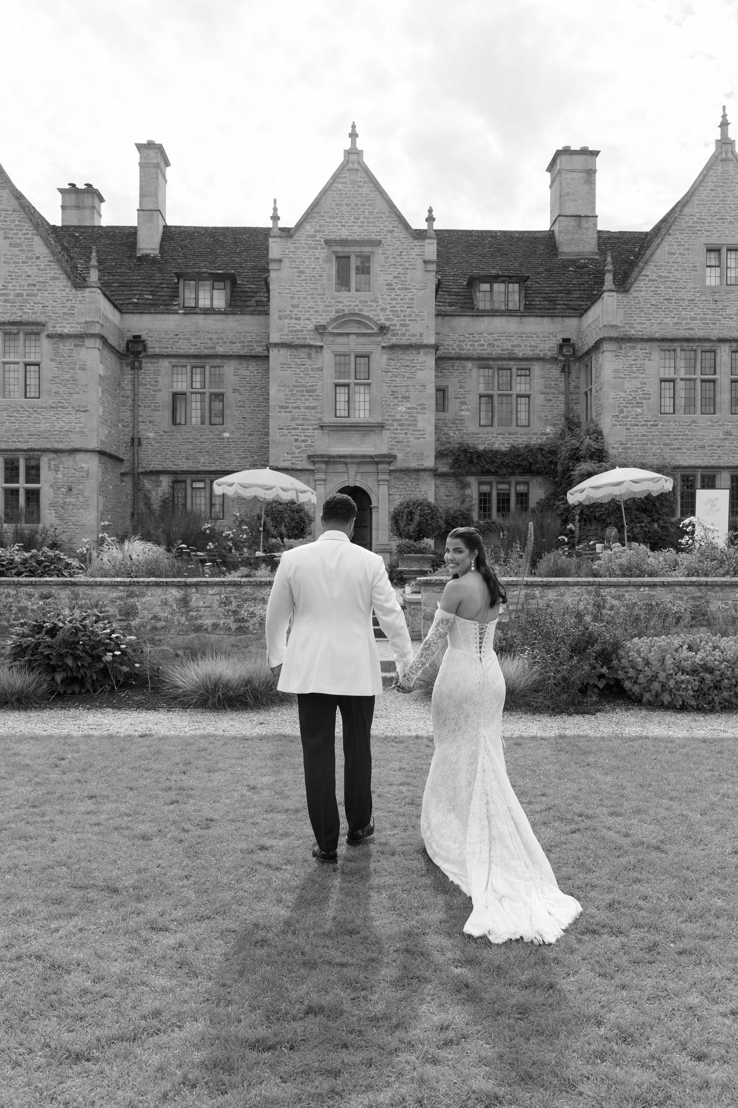 Black and white photo of a bride and groom holding hands walking toward a grand manor house.