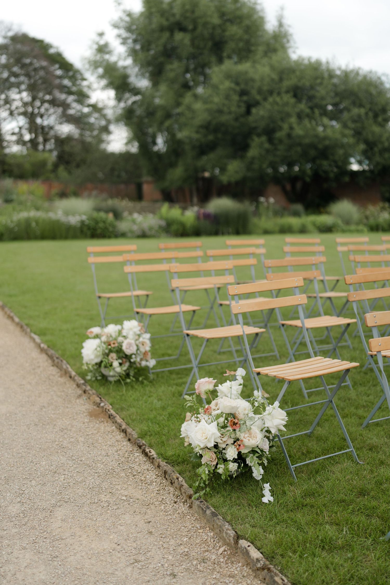 Elegant outdoor wedding ceremony setup with wooden folding chairs on green lawn, adorned with white floral arrangements along the aisle.