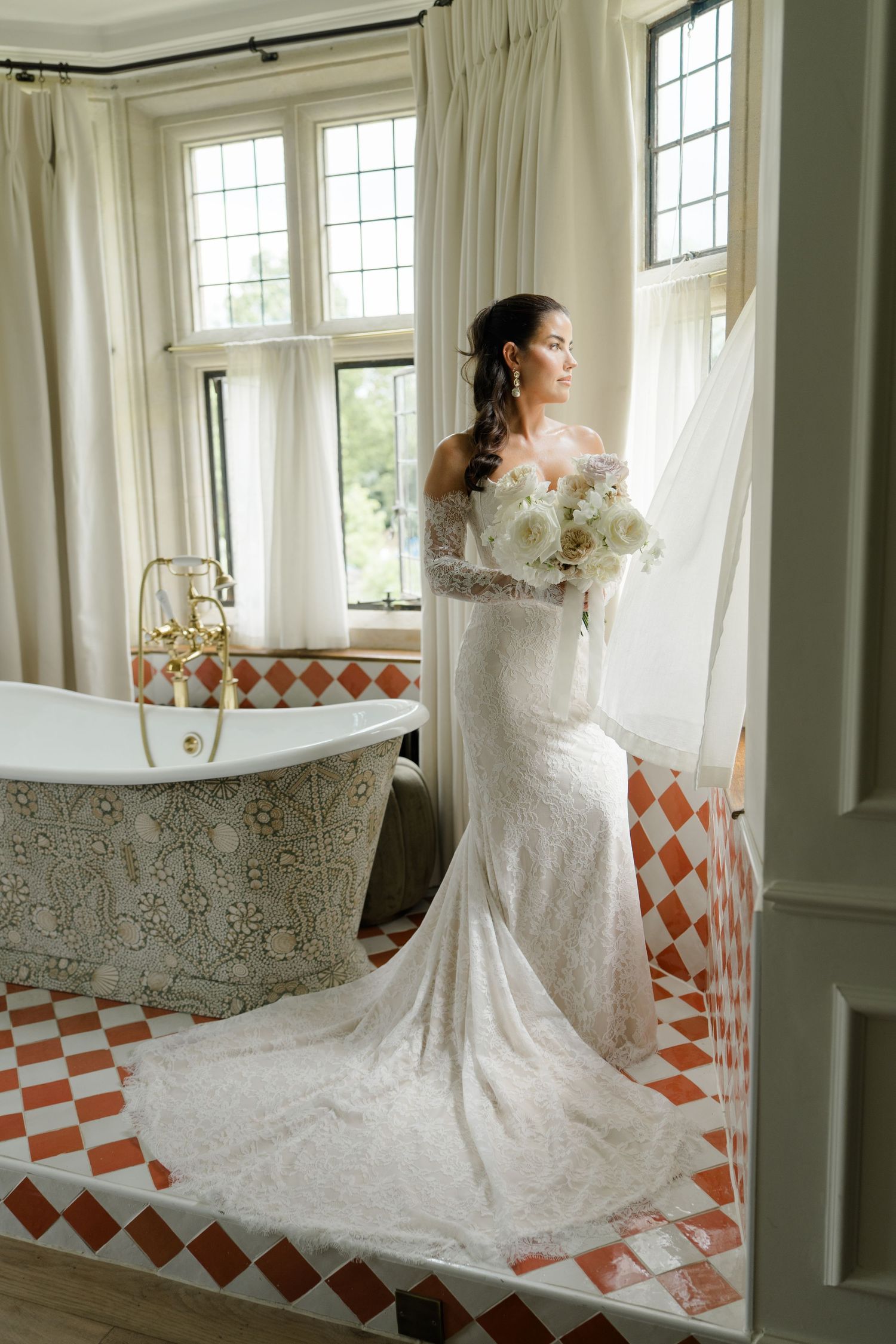 Bride in lace off-shoulder wedding gown holding white bouquet beside clawfoot tub in elegant bathroom.