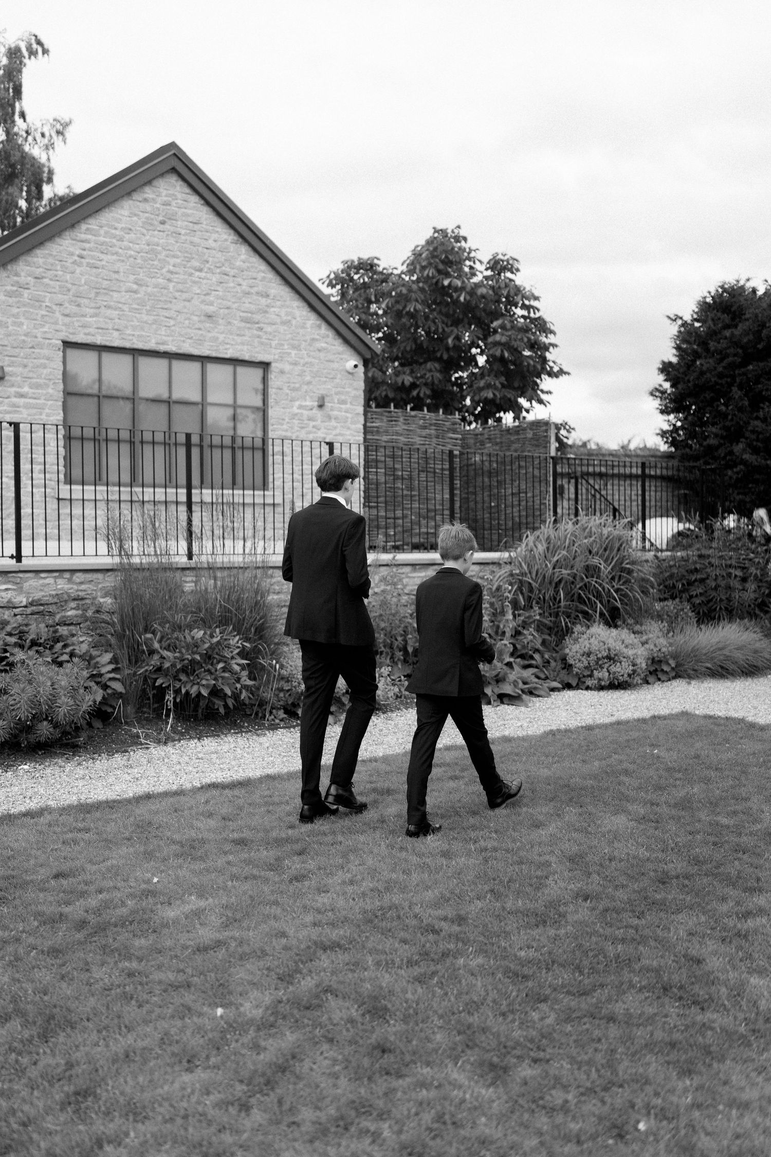 Black and white photo of two boys in suits walking across a garden lawn near a building.