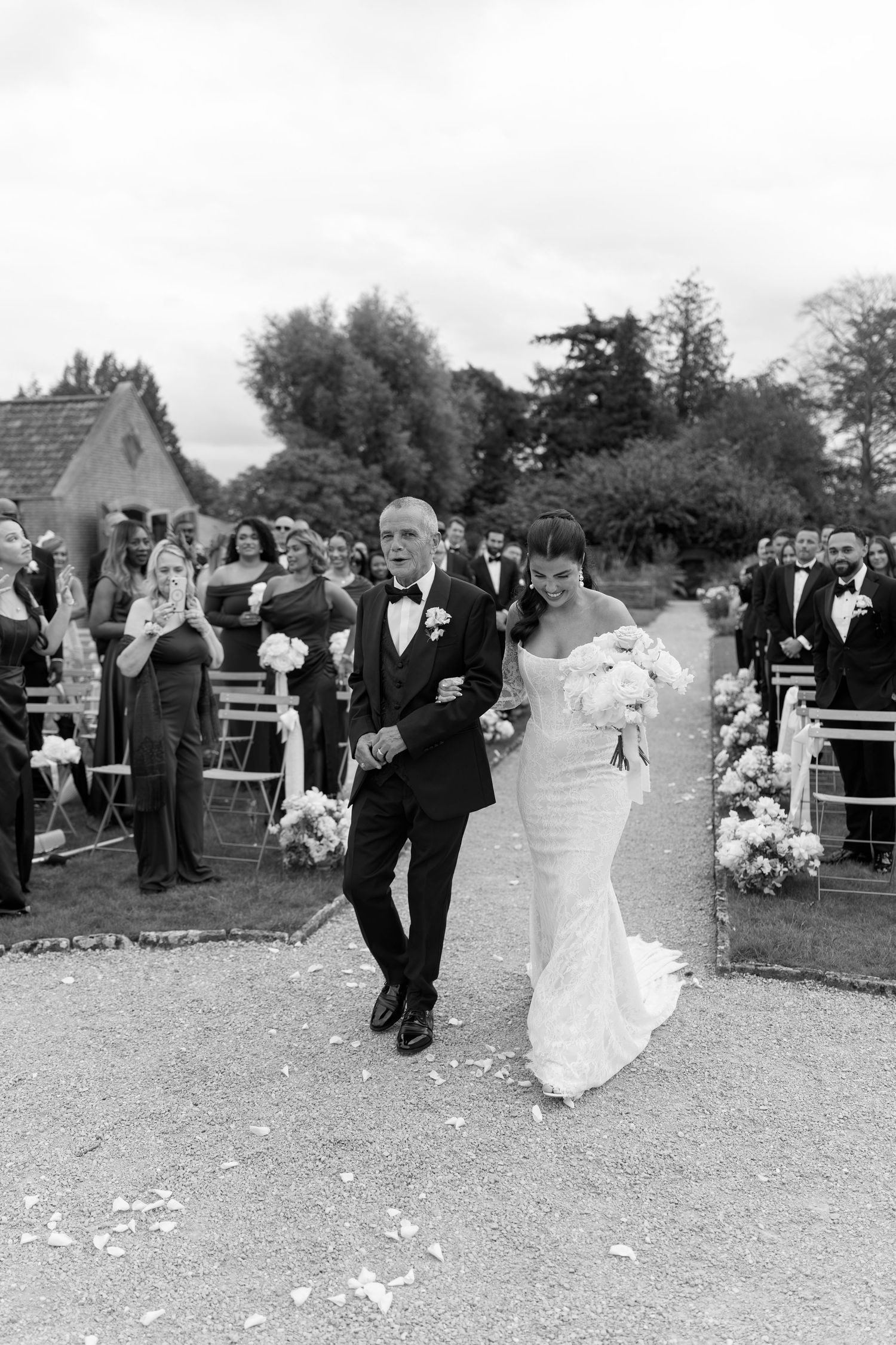 Black and white photo of bride and father walking down outdoor wedding ceremony aisle.