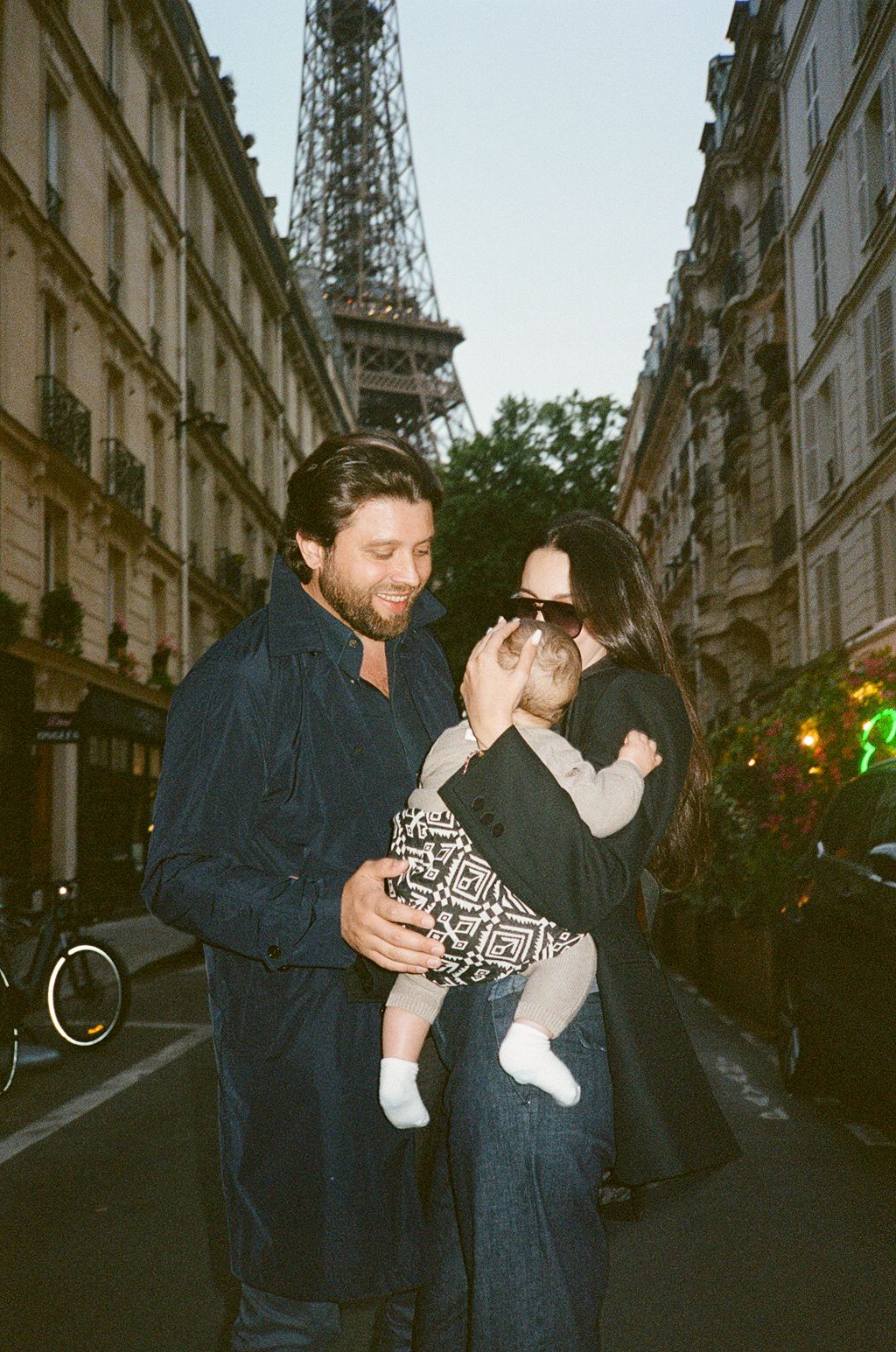 Couple with baby in front of Eiffel Tower on Paris street at dusk.