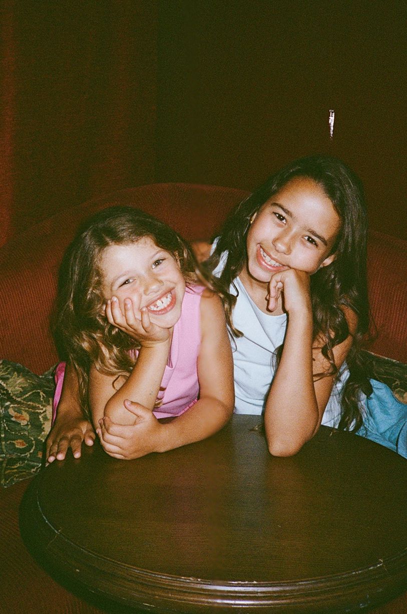 Two young girls smiling and posing together at a wooden table in a warmly lit room.